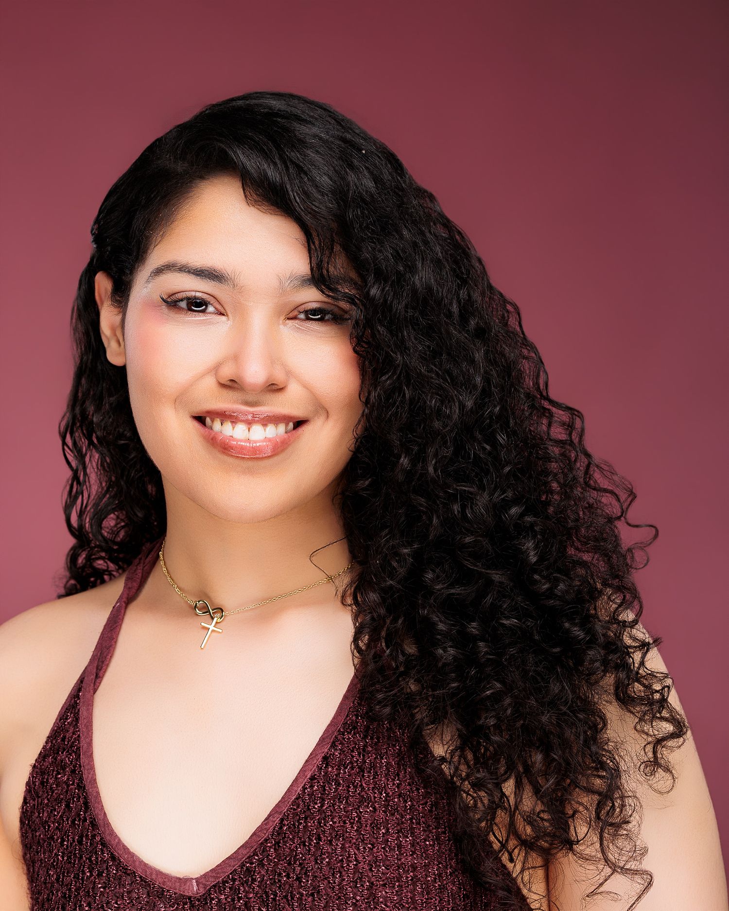 Young woman with long curly black hair smiling, wearing a burgundy top and gold necklace against a mauve background.