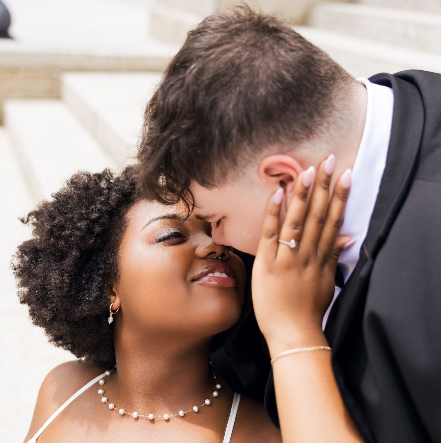 Interracial couple sharing a romantic kiss, bride in white dress with pearl necklace and engagement ring visible.