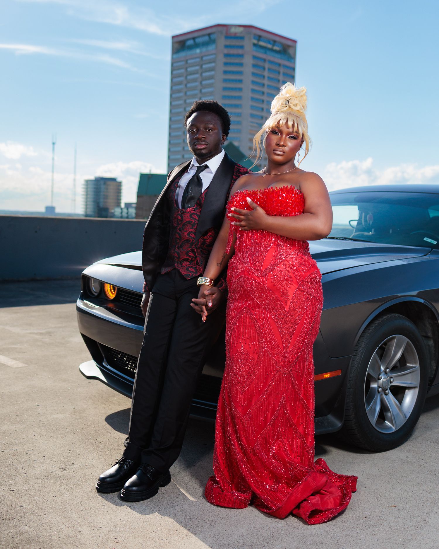 Couple in formal prom attire, man in black tuxedo and woman in red sequin gown, posing by a Dodge Challenger.