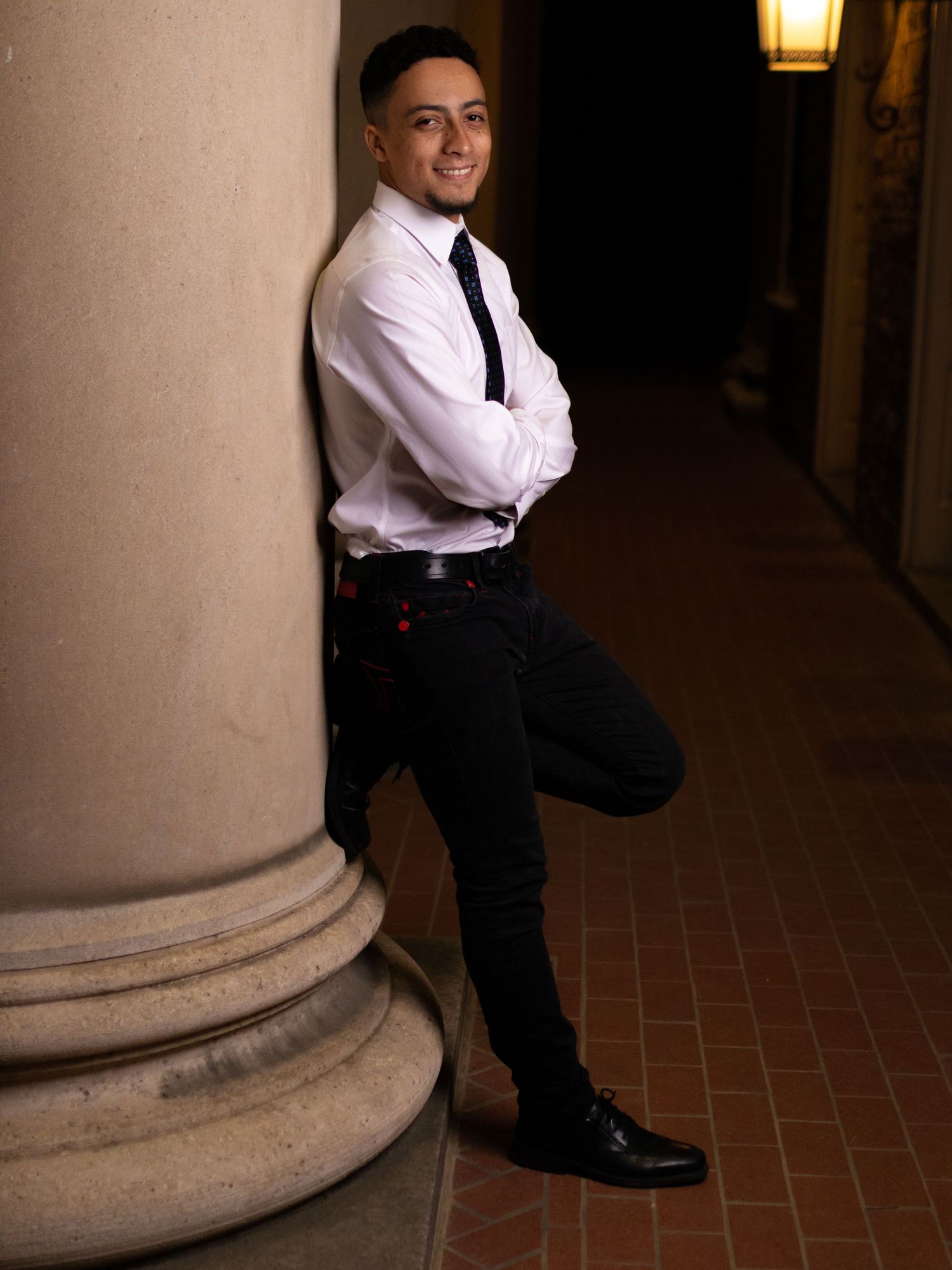 Professional portrait of graduate in formal attire leaning against stone column with warm lighting.