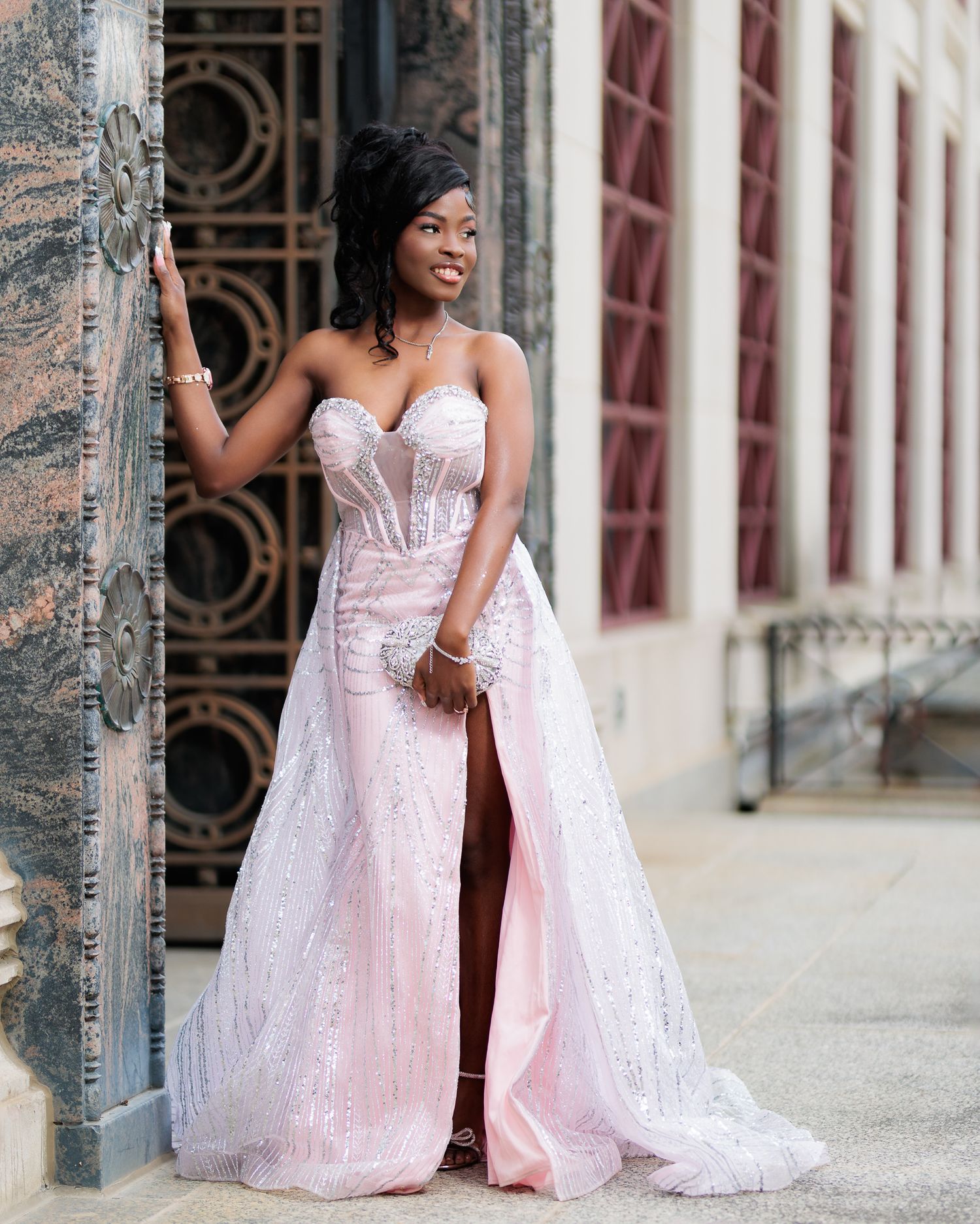 Young woman in a pink sequined strapless gown with a thigh-high slit poses elegantly by an ornate door.