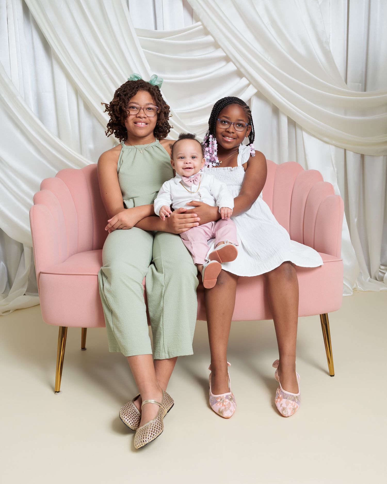 Two girls and a baby boy pose together on a pink velvet chair in a bright studio setting.