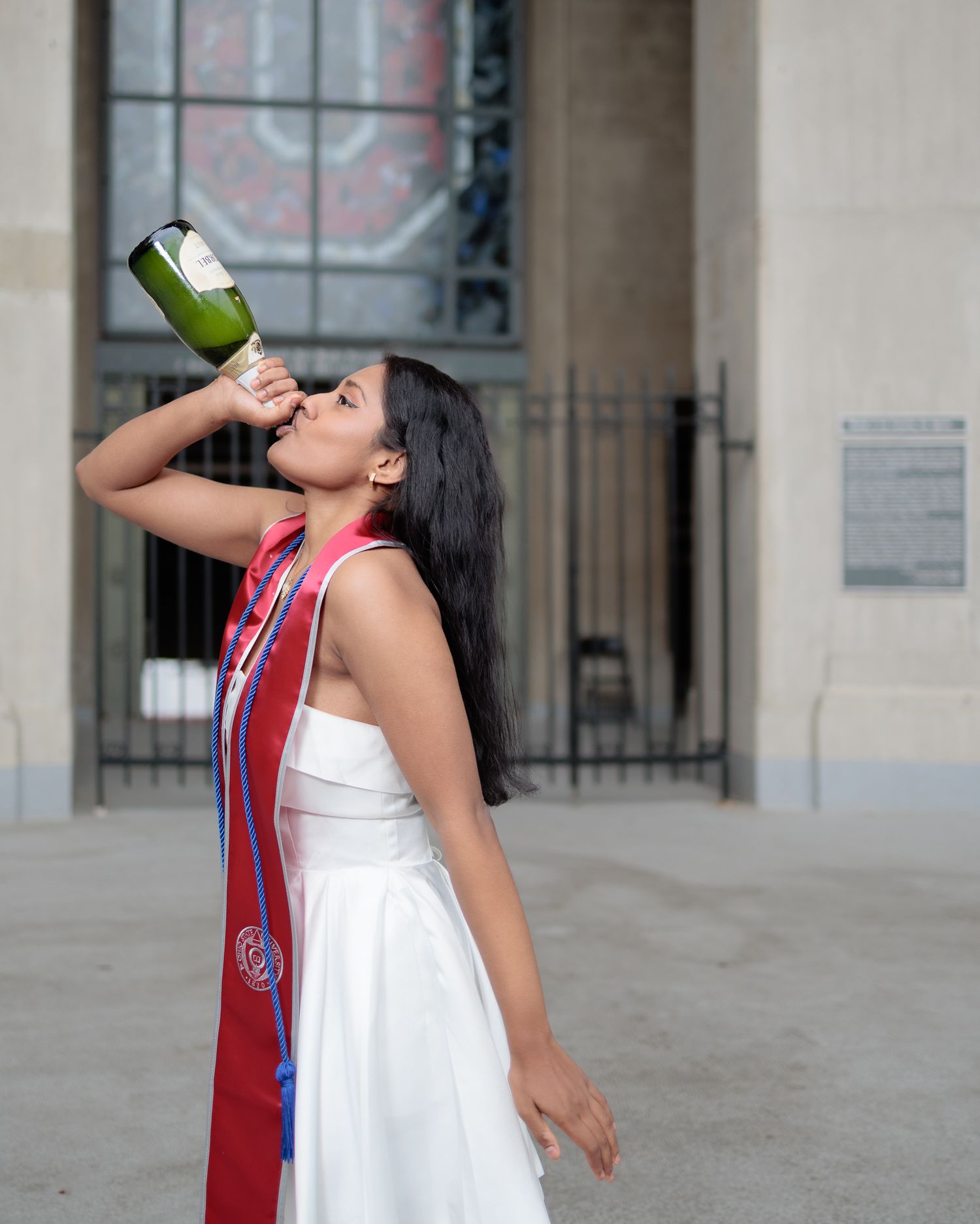 Graduate in white dress and red sash celebrates outdoors by drinking champagne from bottle.