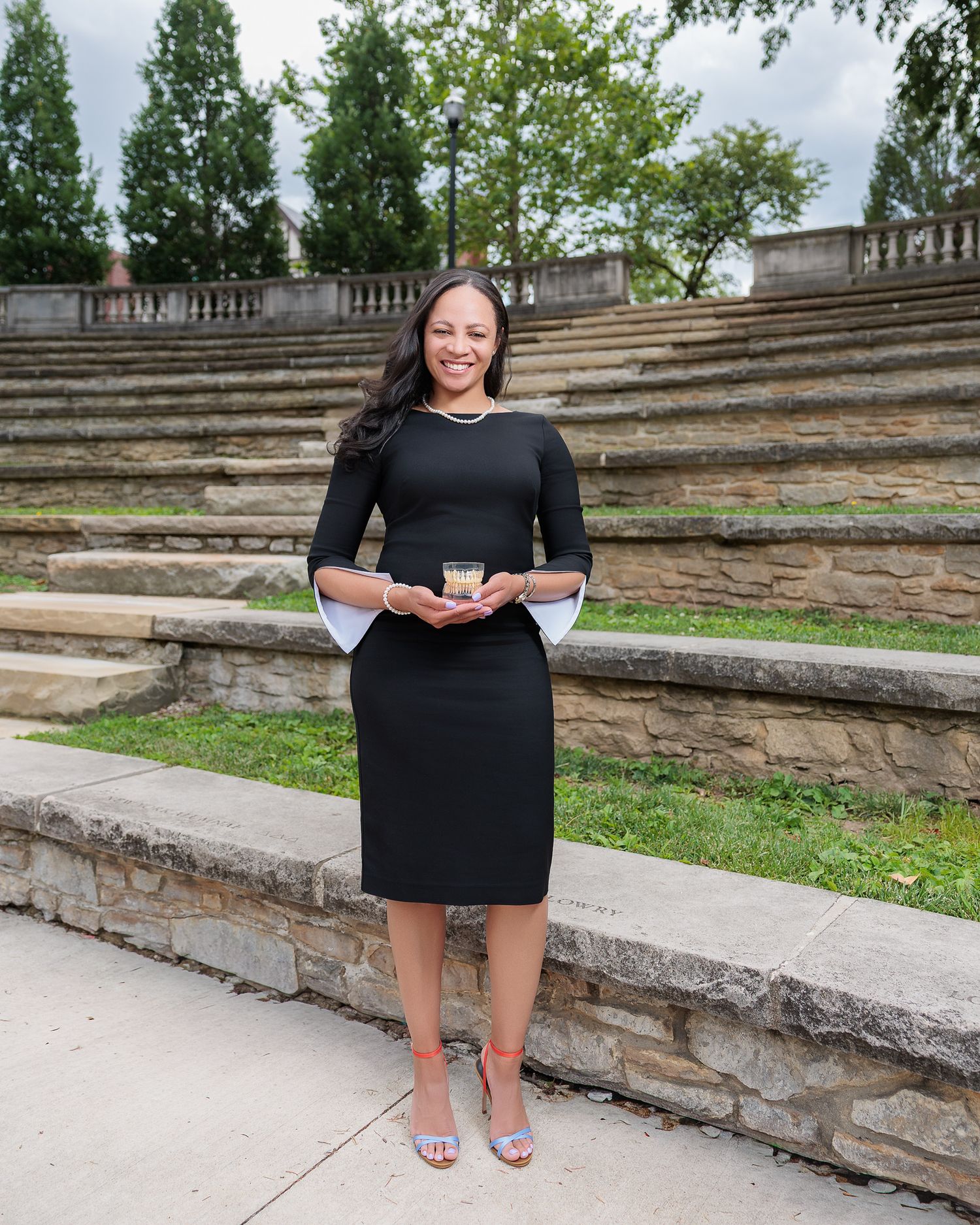 Woman in black dress holding award stands before stone amphitheater steps in outdoor setting.