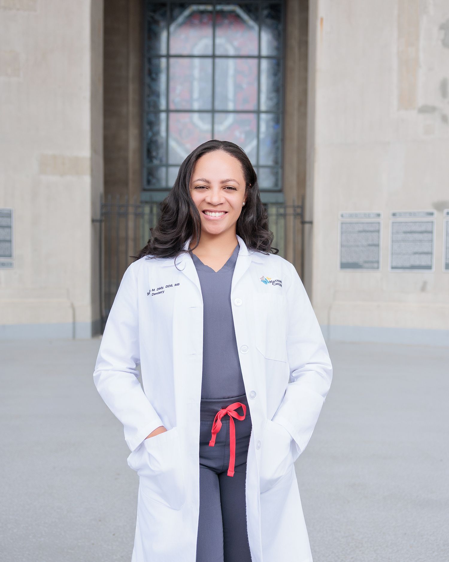 A smiling female doctor in a white lab coat and gray scrubs stands confidently outside a stone building.