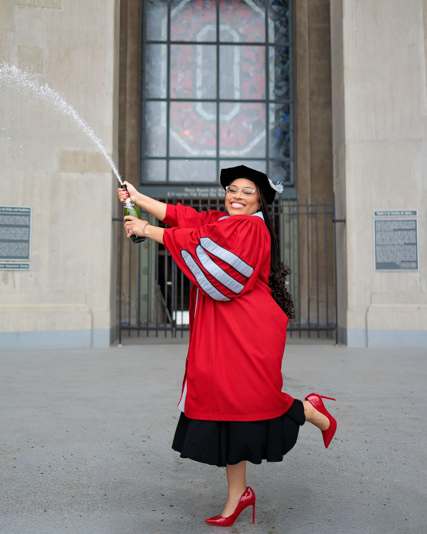 Graduate in red doctoral gown and cap joyfully sprays champagne outside a university building.