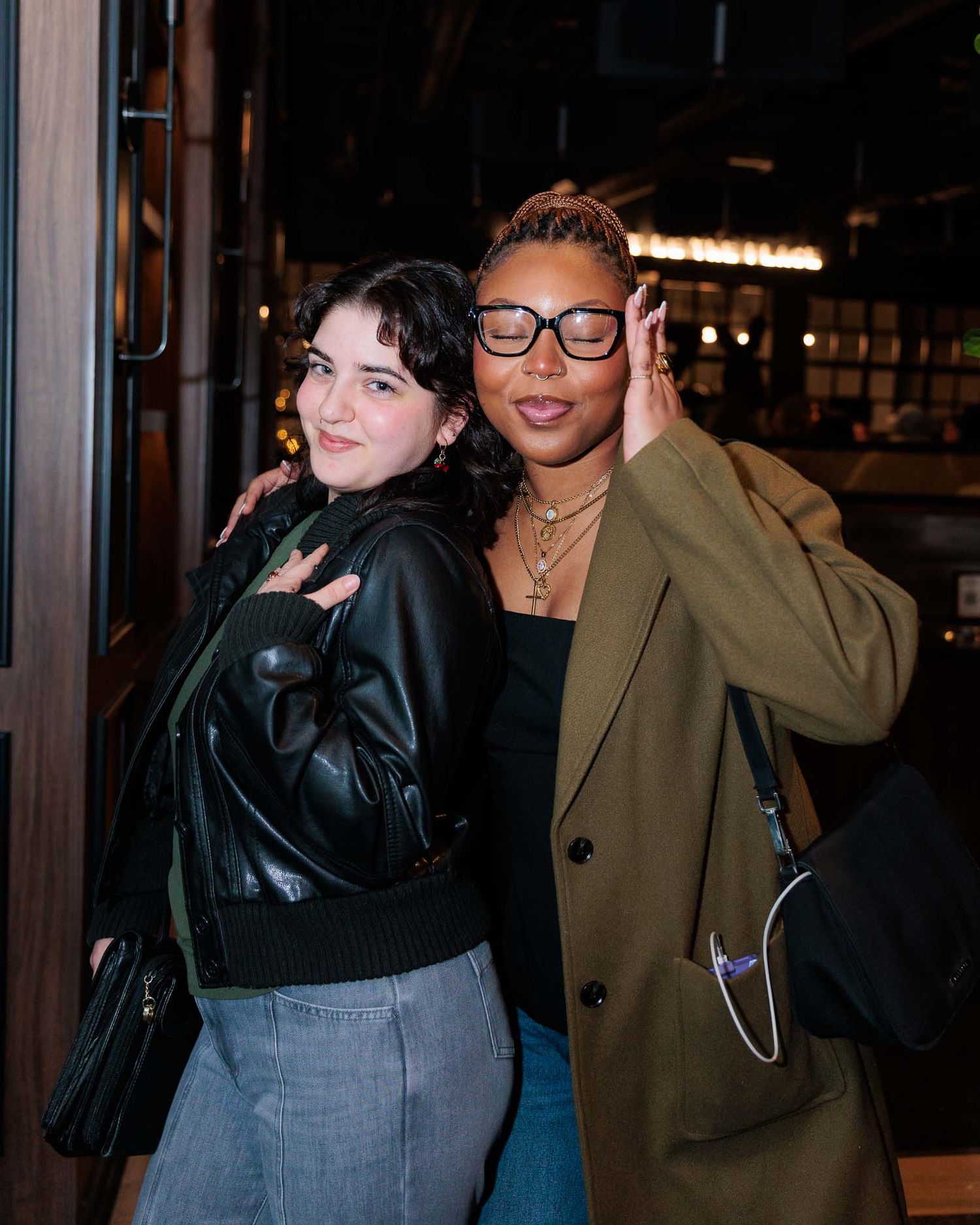 Two people pose together in front of glass windows at night, one wearing a tan coat and one in black.