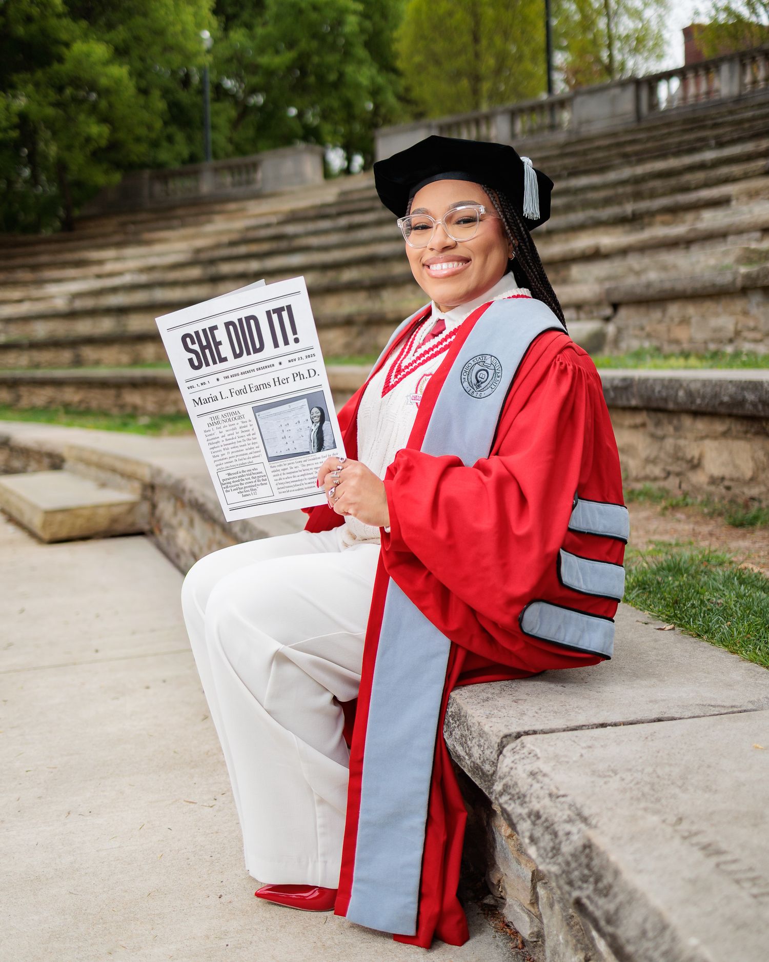 Woman in red doctoral graduation regalia holds newspaper reading She Did It while sitting on stone steps outdoors.