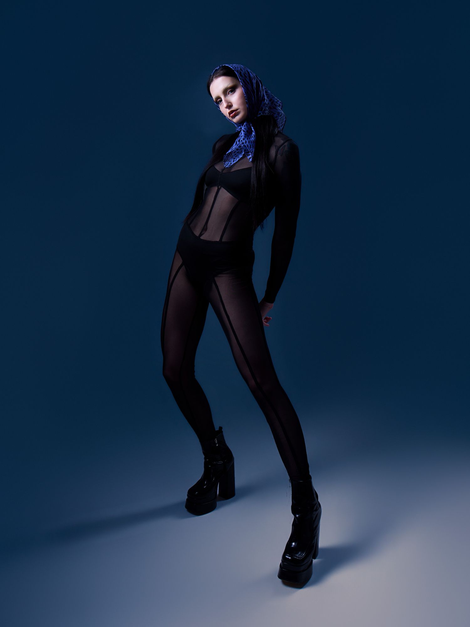 Model in black sheer bodysuit with platform boots and blue scarf poses against dark blue background.