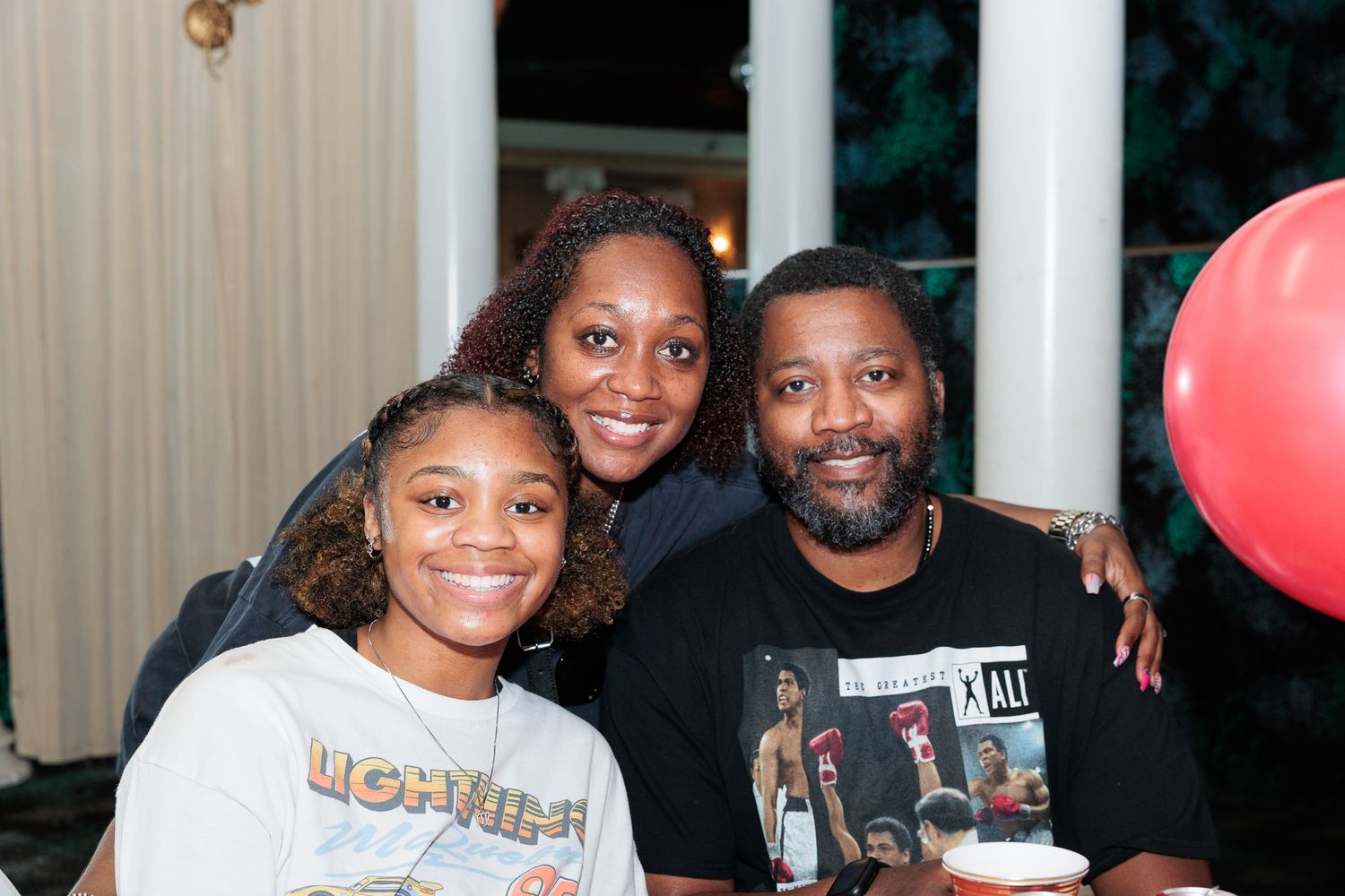 Family of three posing together at a celebration with red balloons in the background.