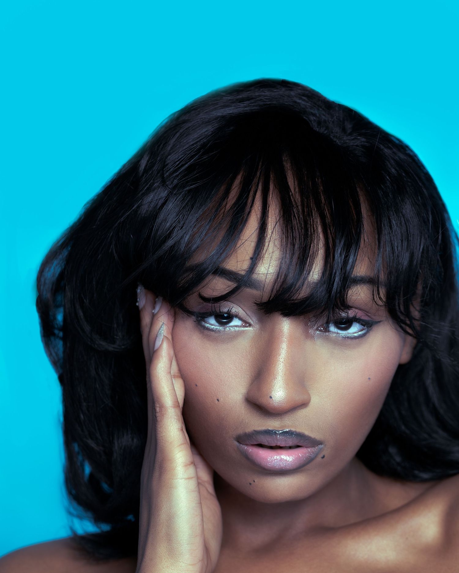 A young Black woman with black wavy hair and bangs rests her hand on her face against a cyan background.