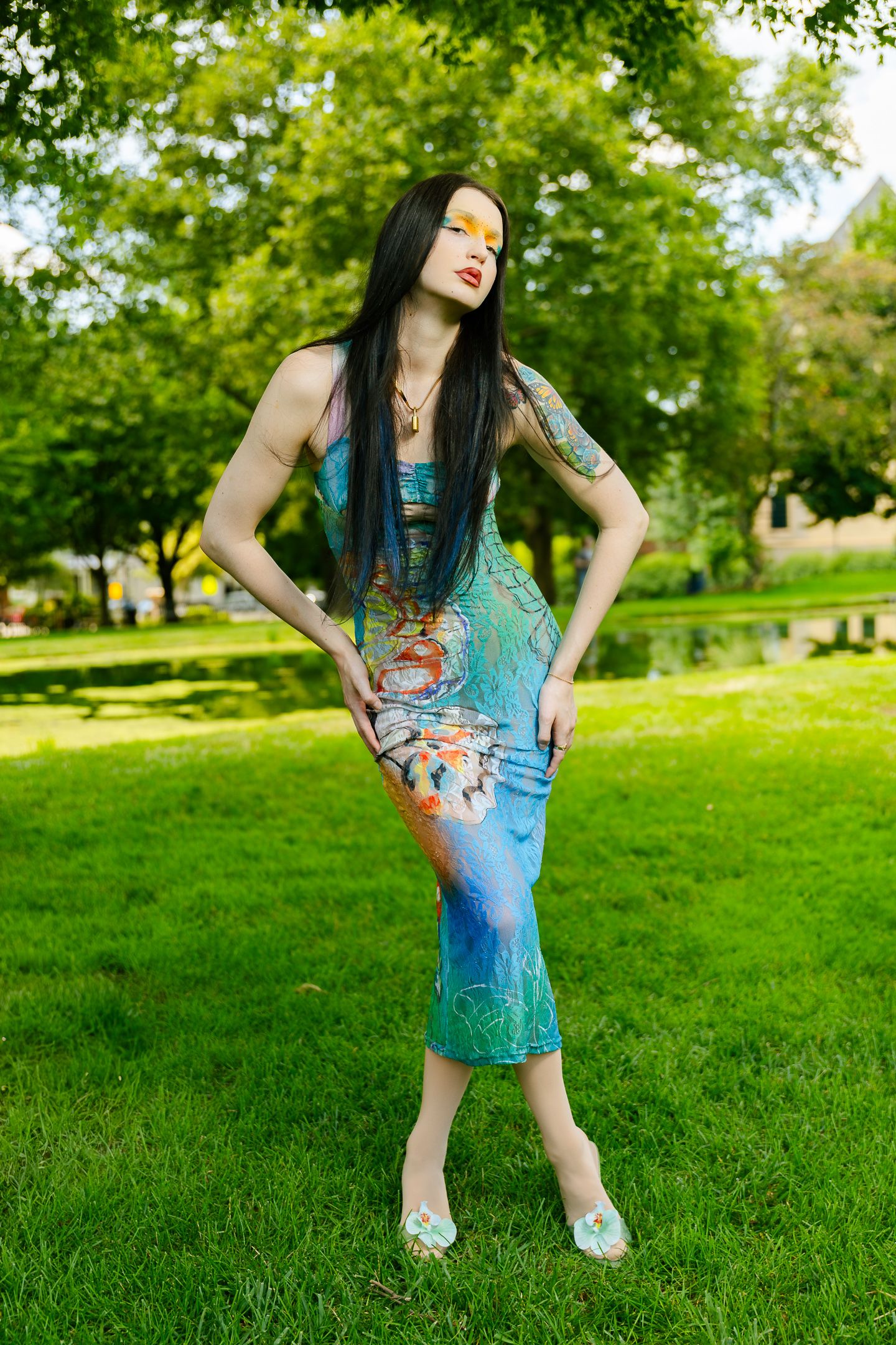 Tattooed woman in vibrant aquatic maxi dress posing dynamically on green grass in outdoor park.