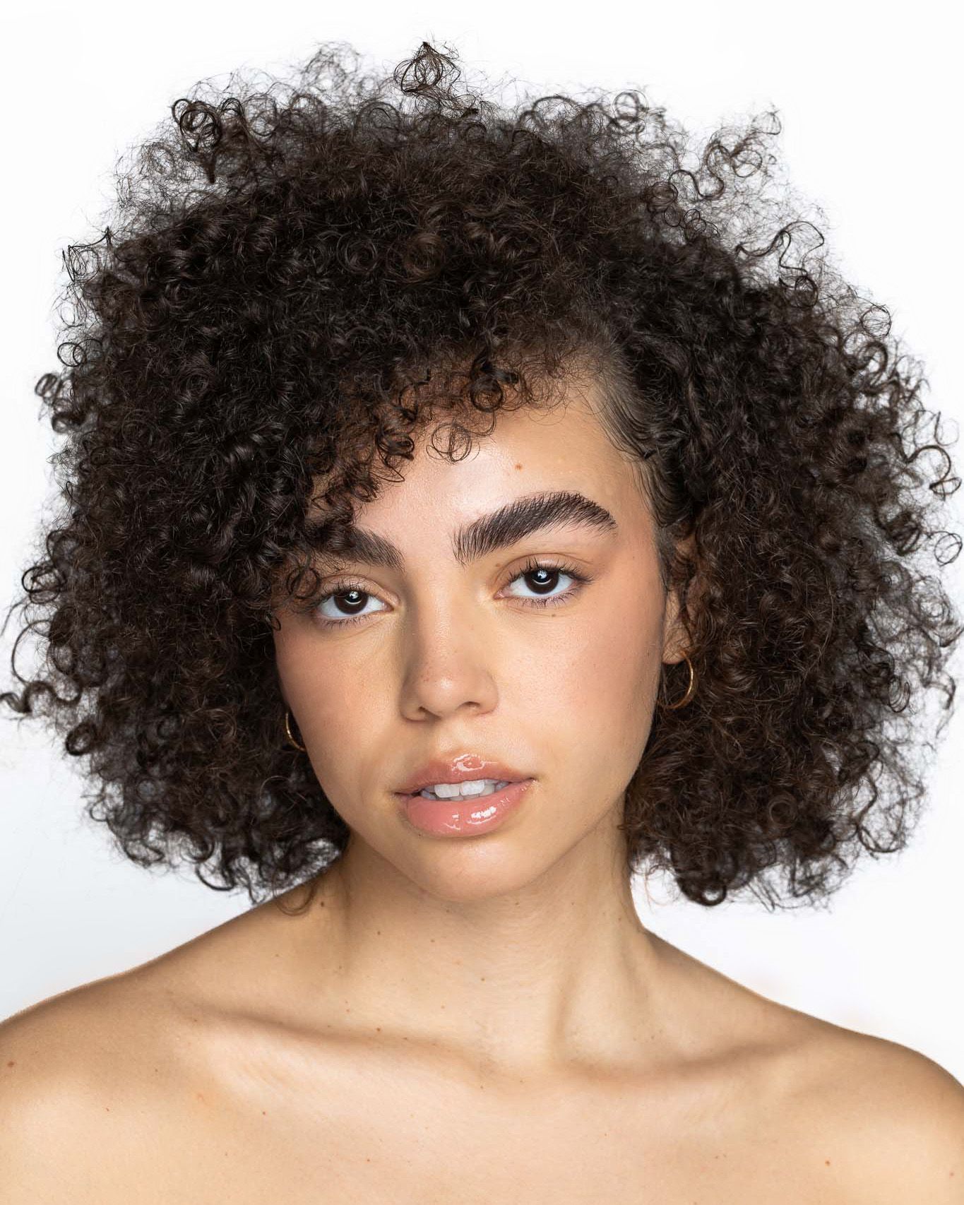 Portrait against white background featuring voluminous natural dark curly hair, bare shoulders, and glowing skin.
