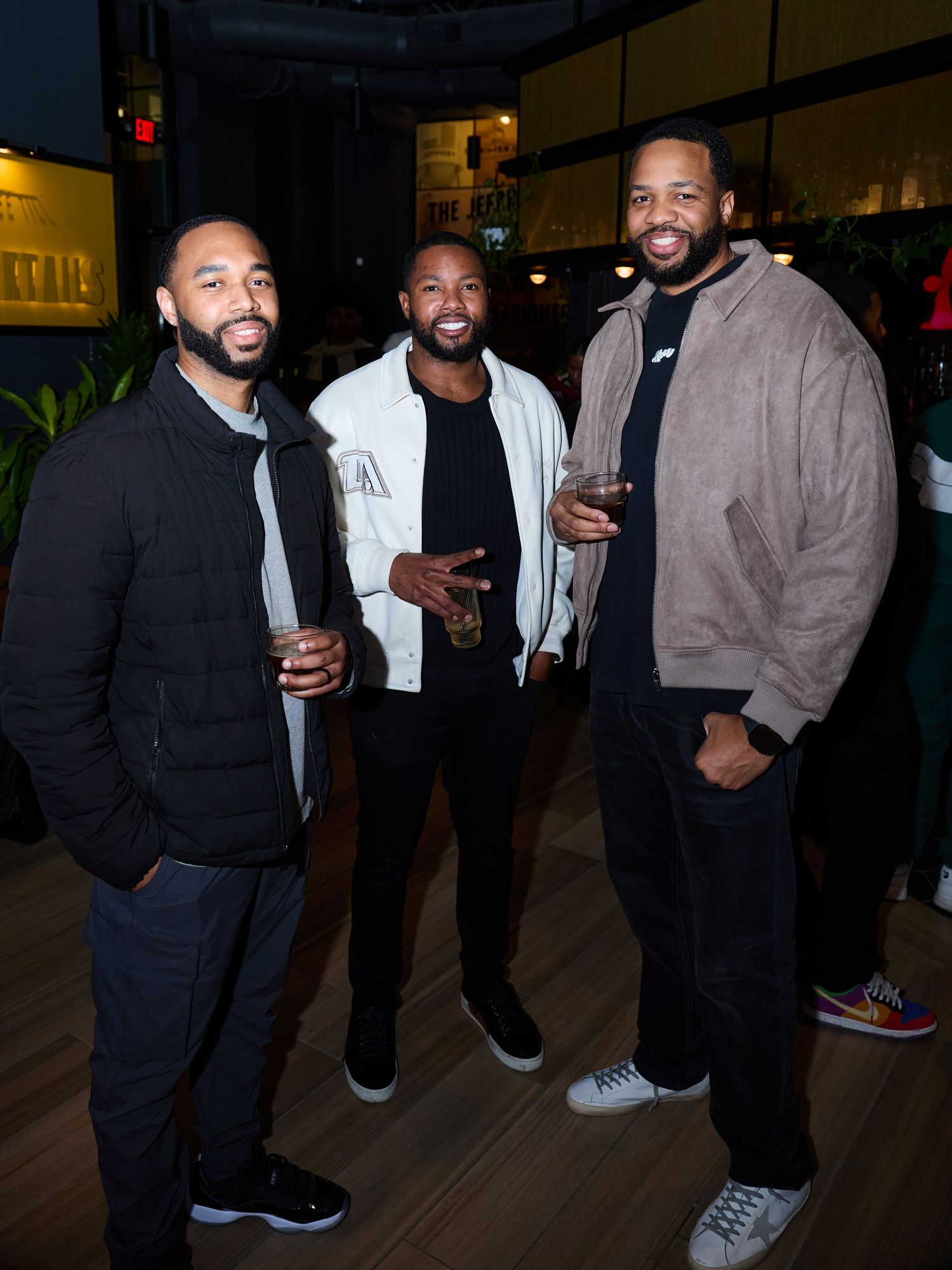 Three friends enjoying drinks and conversation at a dimly lit bar in casual evening attire.