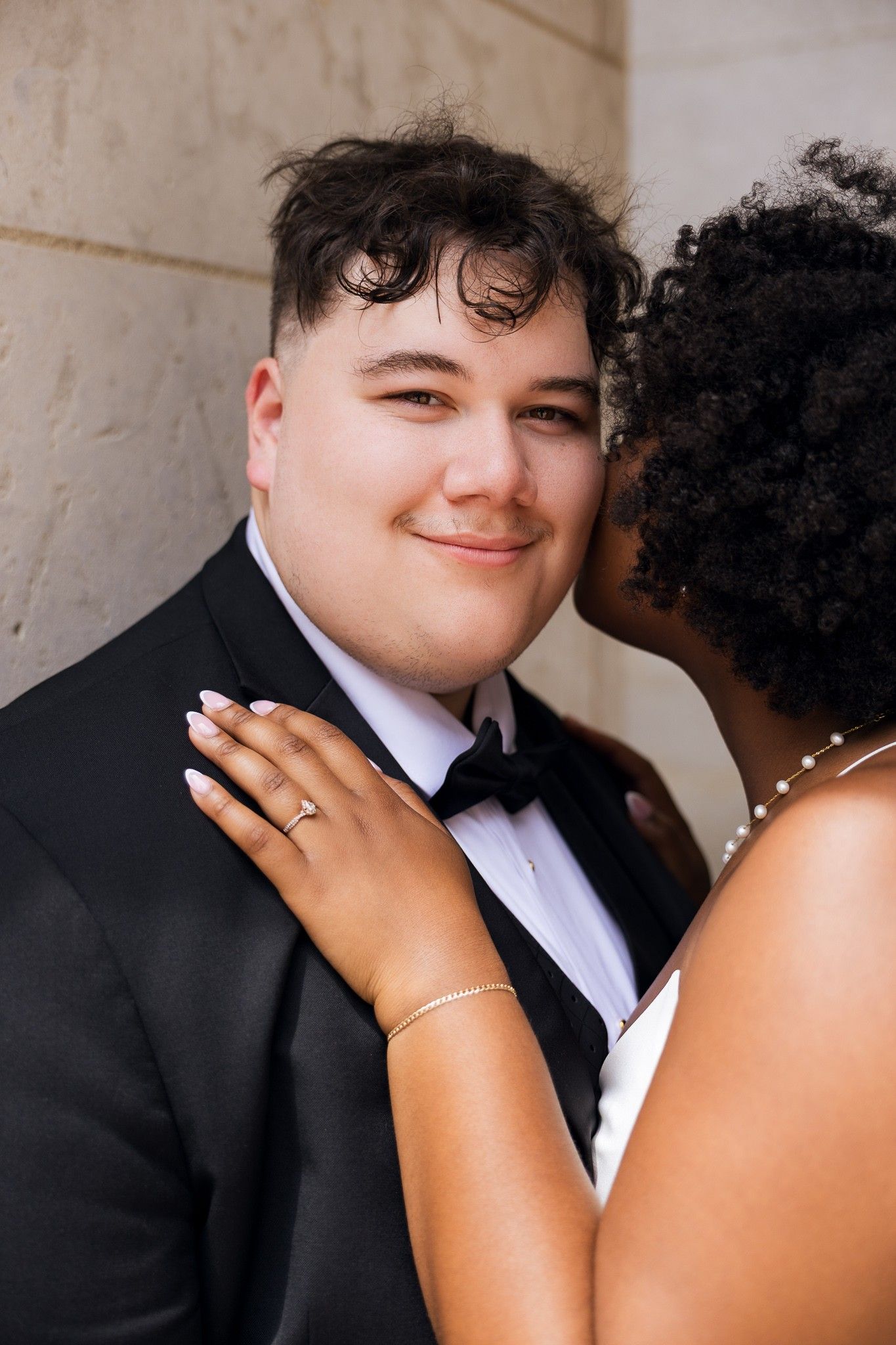 A groom in a black tuxedo smiles as his bride whispers in his ear, her hand resting on his chest.