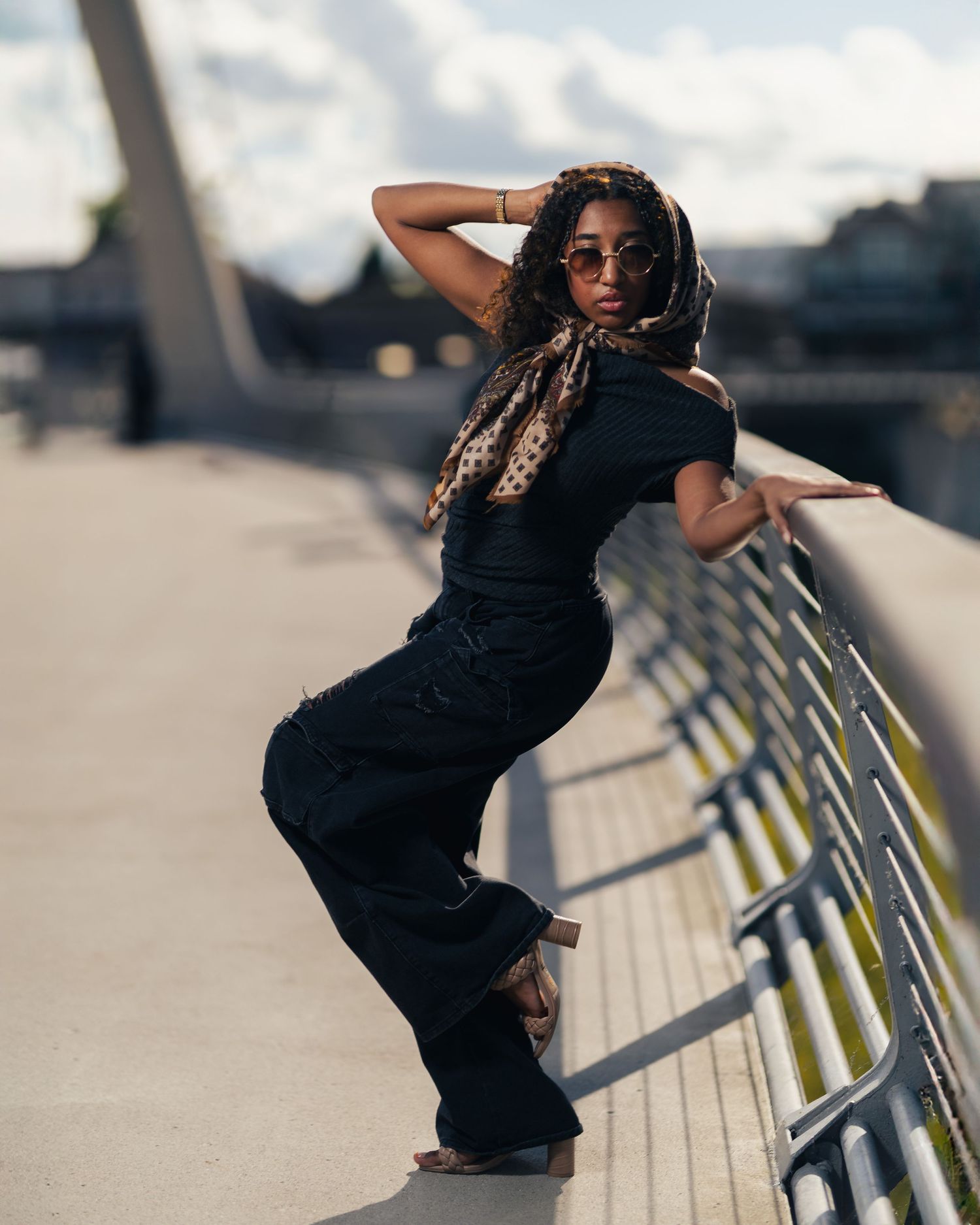 A stylish woman in all-black outfit poses confidently on a bridge railing, wearing sunglasses and a leopard print headscarf.