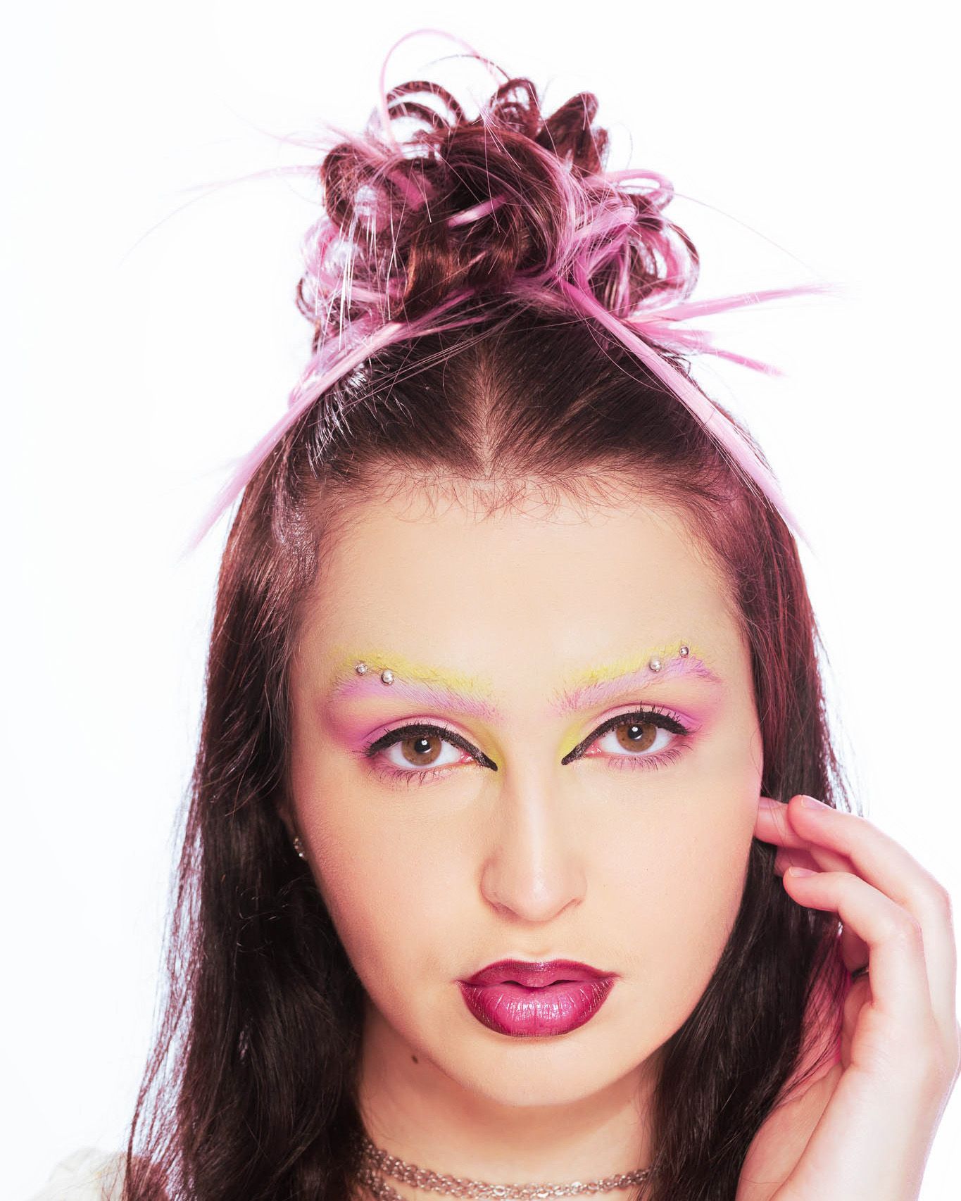 Woman with bold pink and yellow eyeshadow, rhinestone brow accents, dark lips, and pink-tipped updo hairstyle.