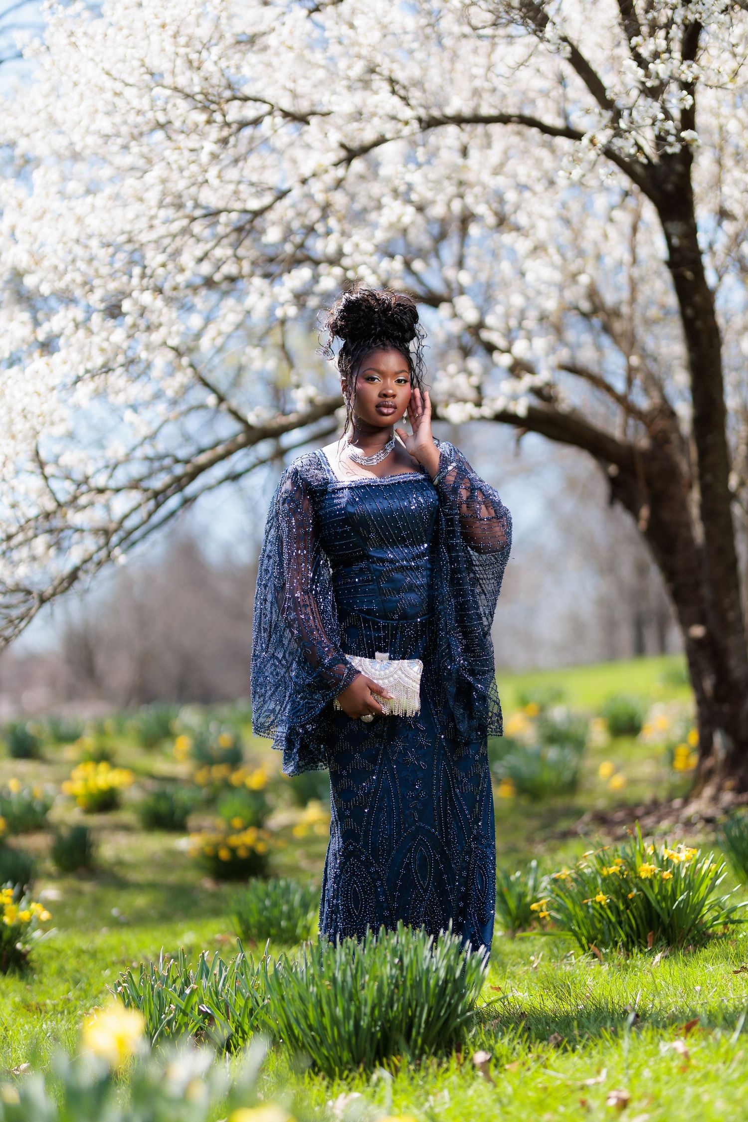 Young Black woman in navy sequin gown poses among spring daffodils and white blossoming trees holding a white clutch.