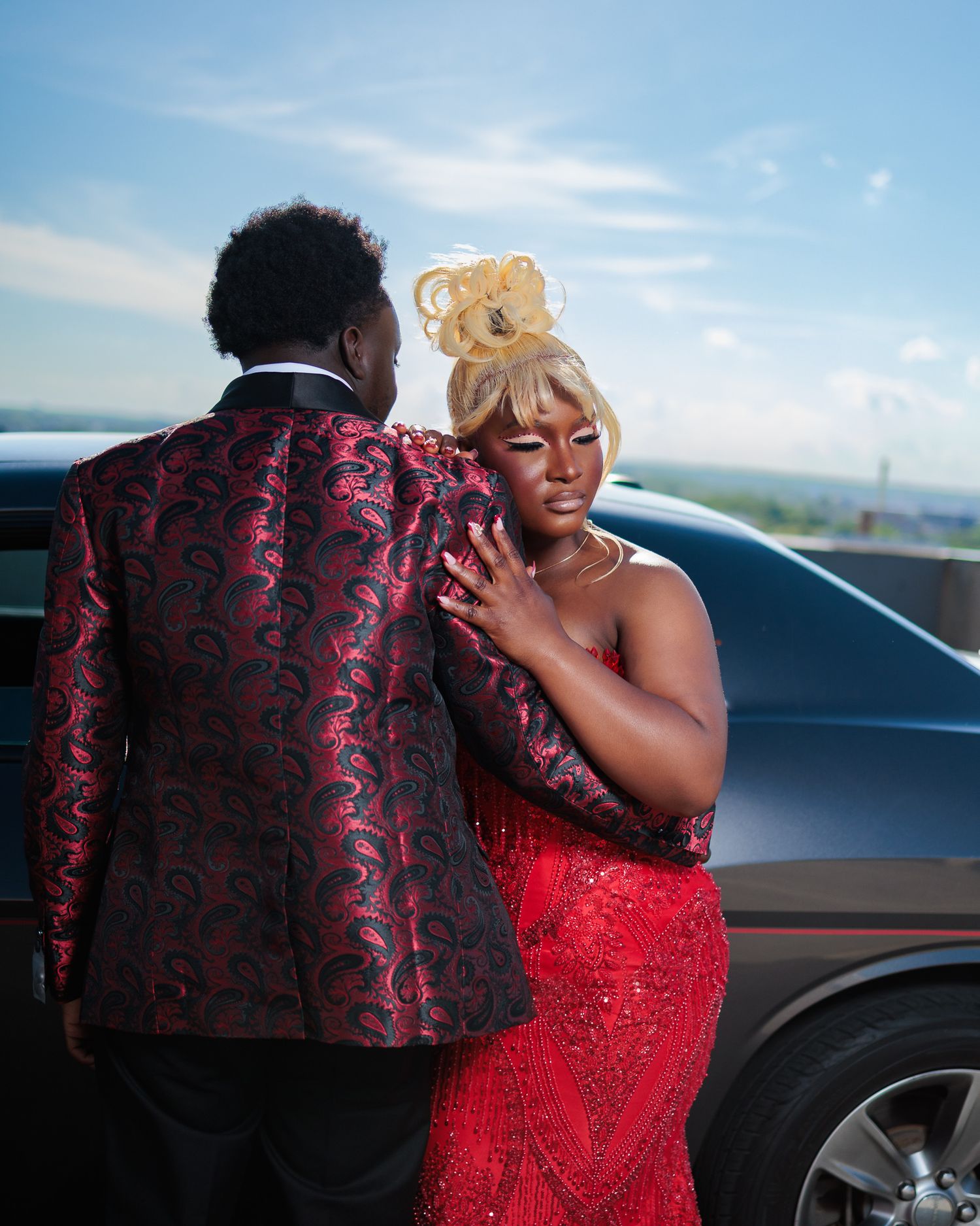 Couple in elegant formal wear, man in red brocade blazer and woman in red sequin dress, posing near a luxury car.