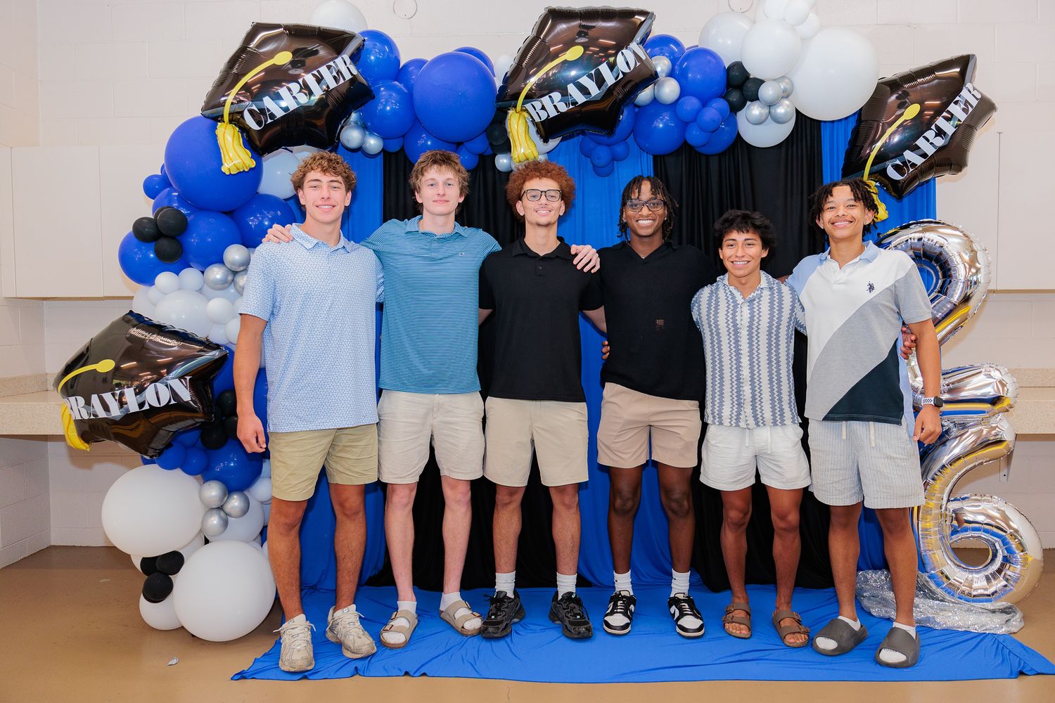 Six teenage boys pose together at a graduation celebration with blue and black balloon arch backdrop.