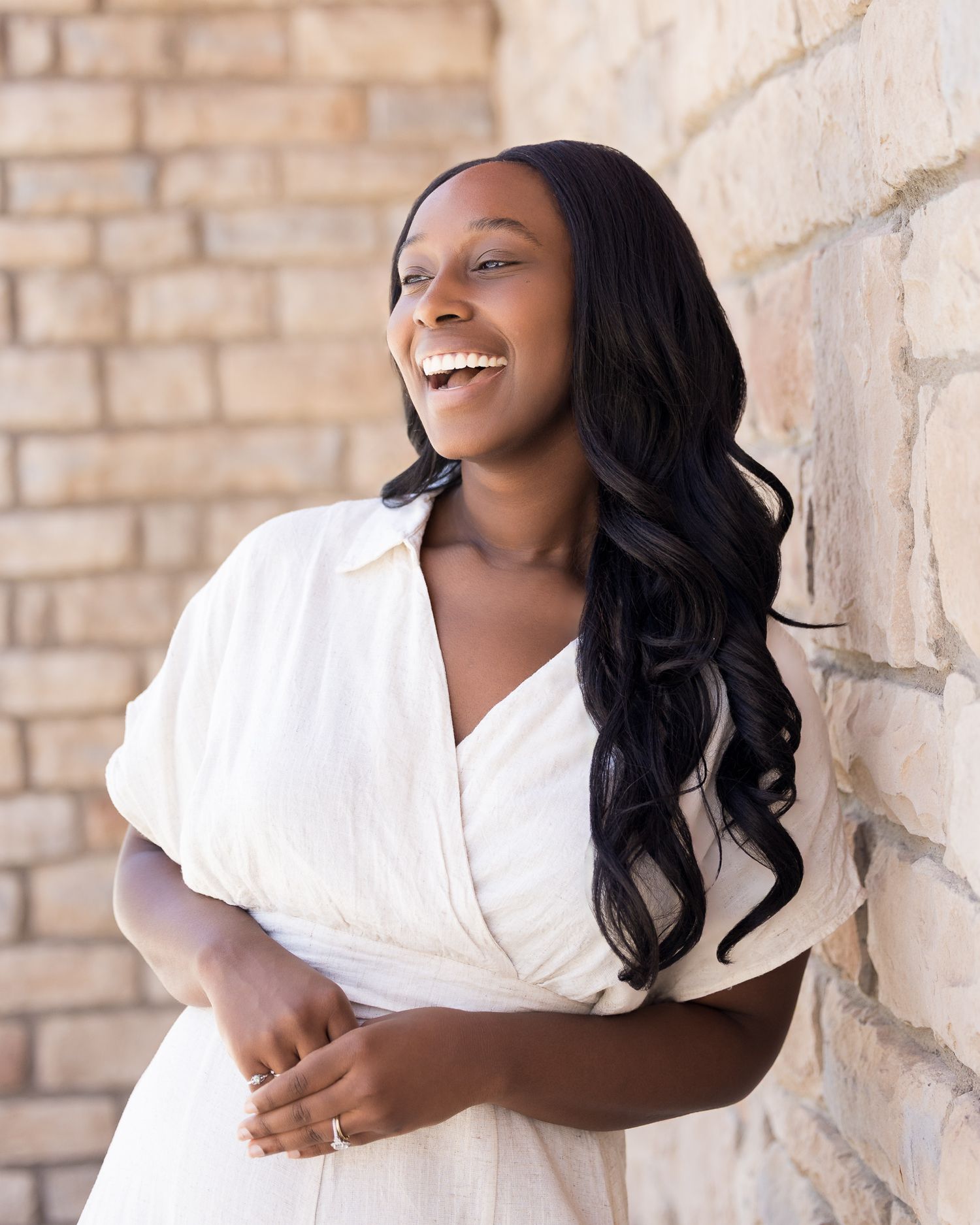 A young Black woman in a white wrap dress laughs joyfully while leaning against a stone wall outdoors.