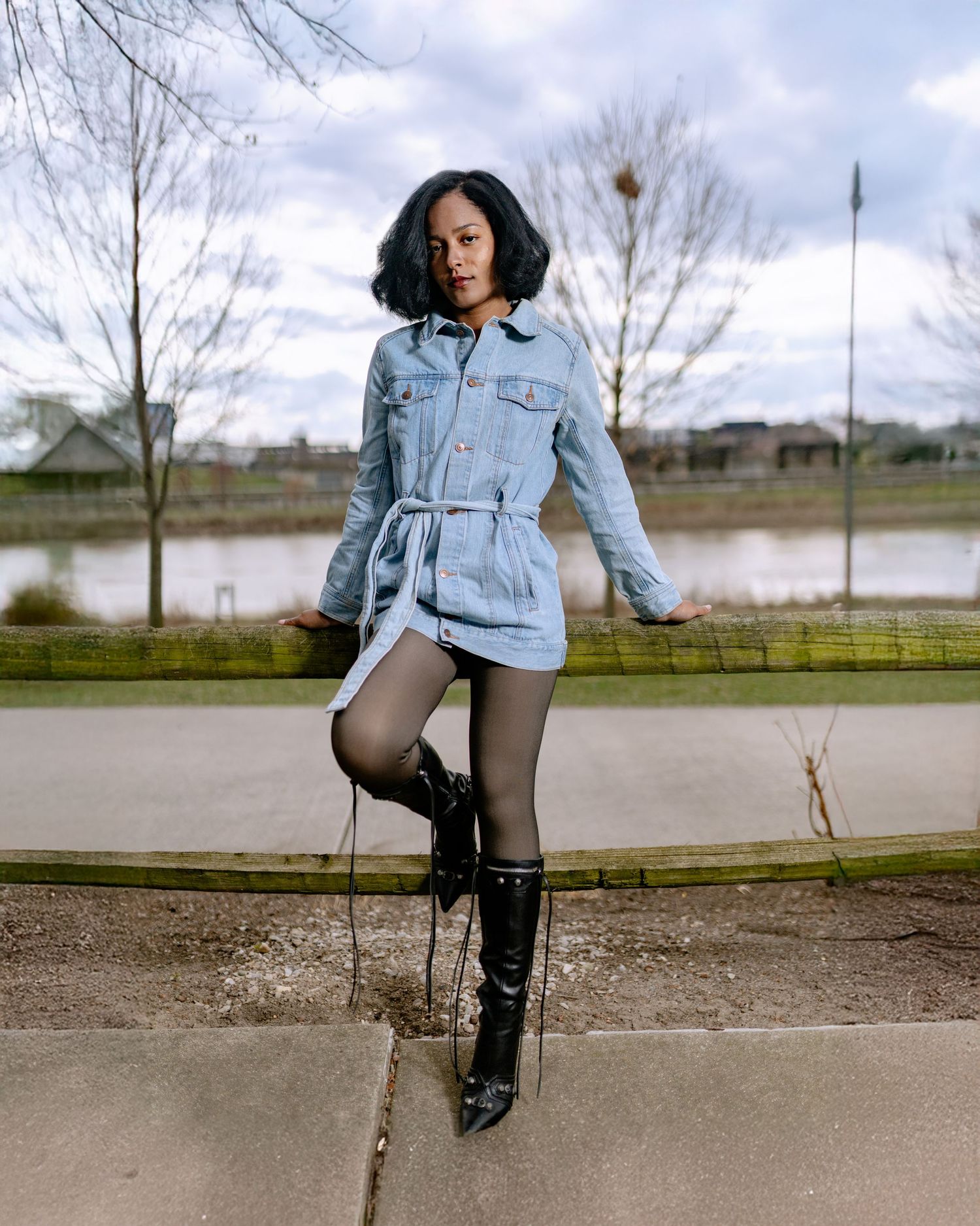 A young woman poses outdoors in a denim mini dress, black tights, and knee-high boots near a park pond.