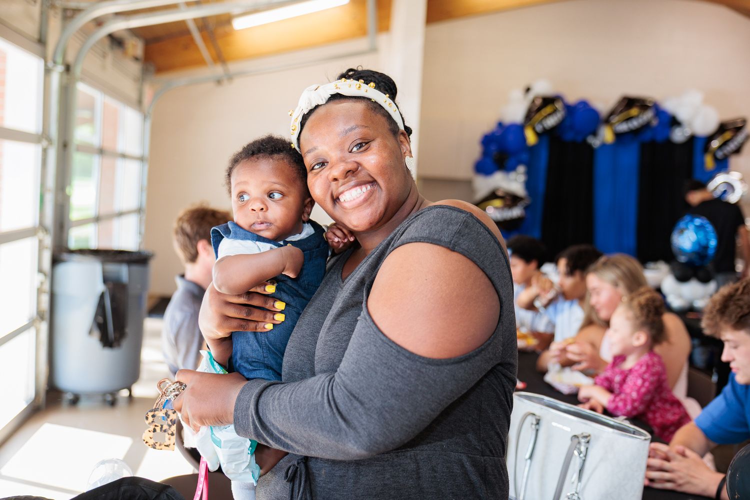 Smiling woman holding a baby at a festive indoor gathering with blue and black decorations.