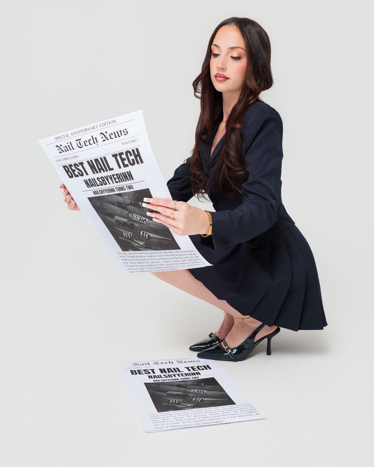 Woman in black dress reading Nail Tech News newspaper, crouching elegantly in heels on white background.