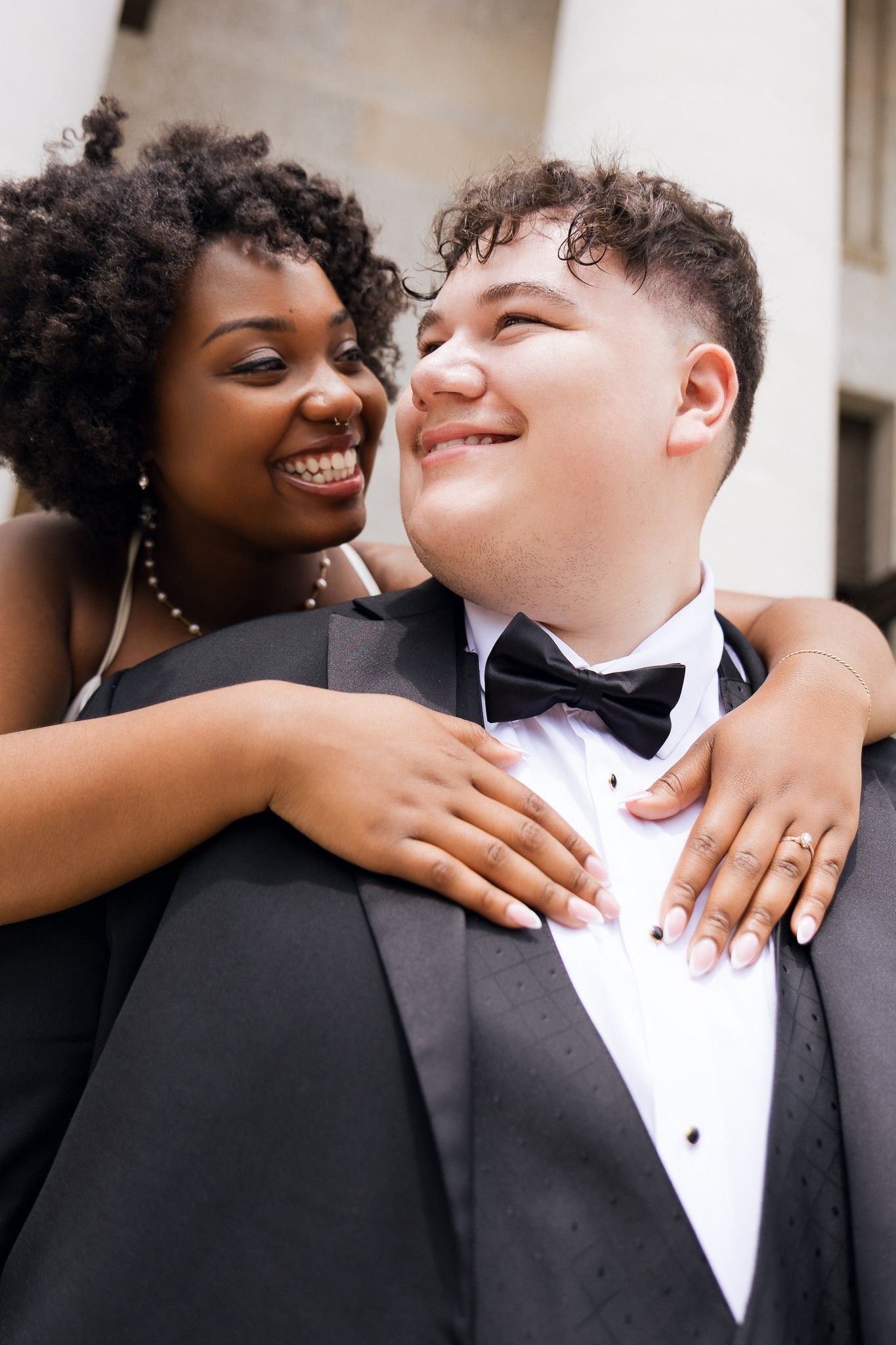 A joyful interracial couple in formal attire, woman in black dress hugging man in tuxedo with bow tie, smiling outdoors.