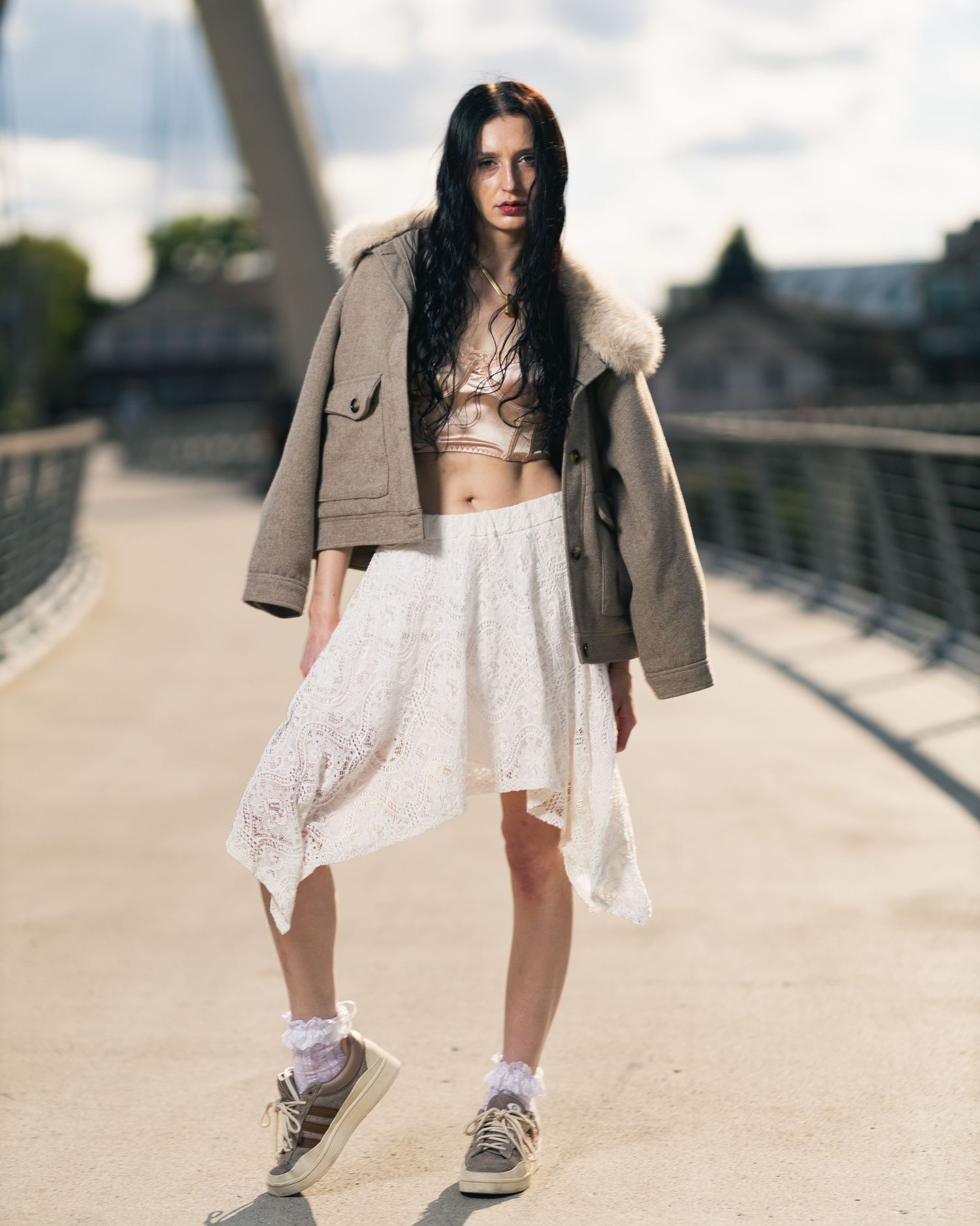 Young woman in olive parka, white lace skirt, and sneakers poses on a bridge in a street fashion editorial.