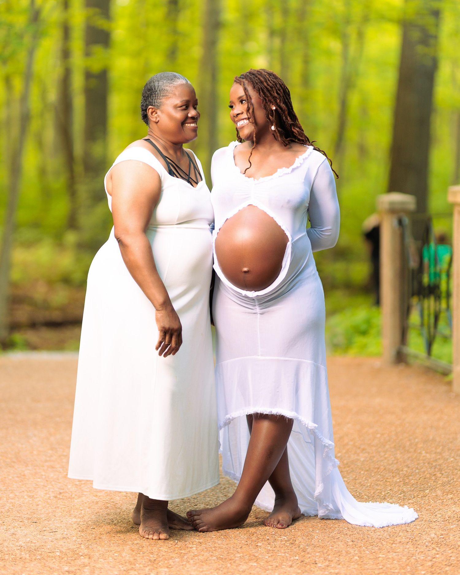 Two women in white dresses pose outdoors, one visibly pregnant, sharing a joyful moment in a lush green forest setting.