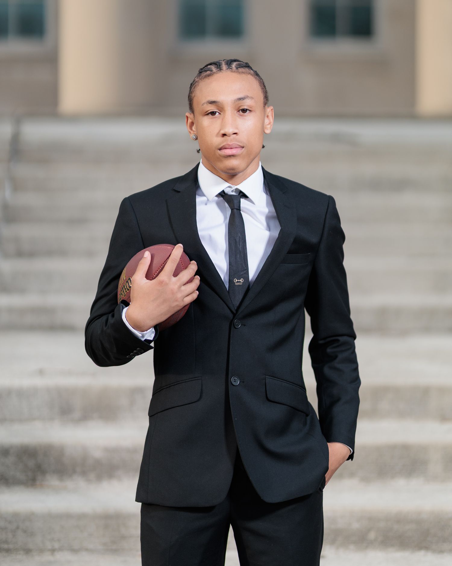 Young man in black suit holding football on steps, formal senior portrait photo.