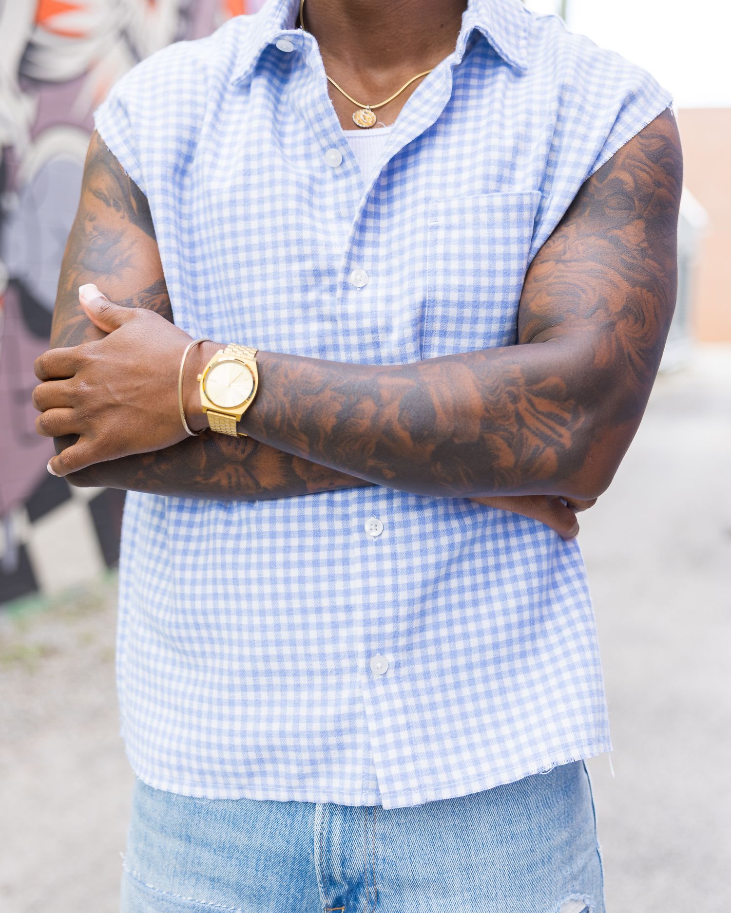 Man with tattoo sleeves wearing blue gingham sleeveless shirt, gold watch, and necklace with arms crossed.