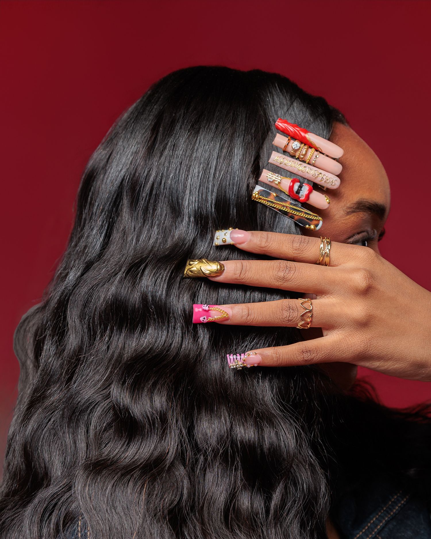 Woman with long wavy black hair showcasing elaborate colorful nail art designs on both hands against red background.