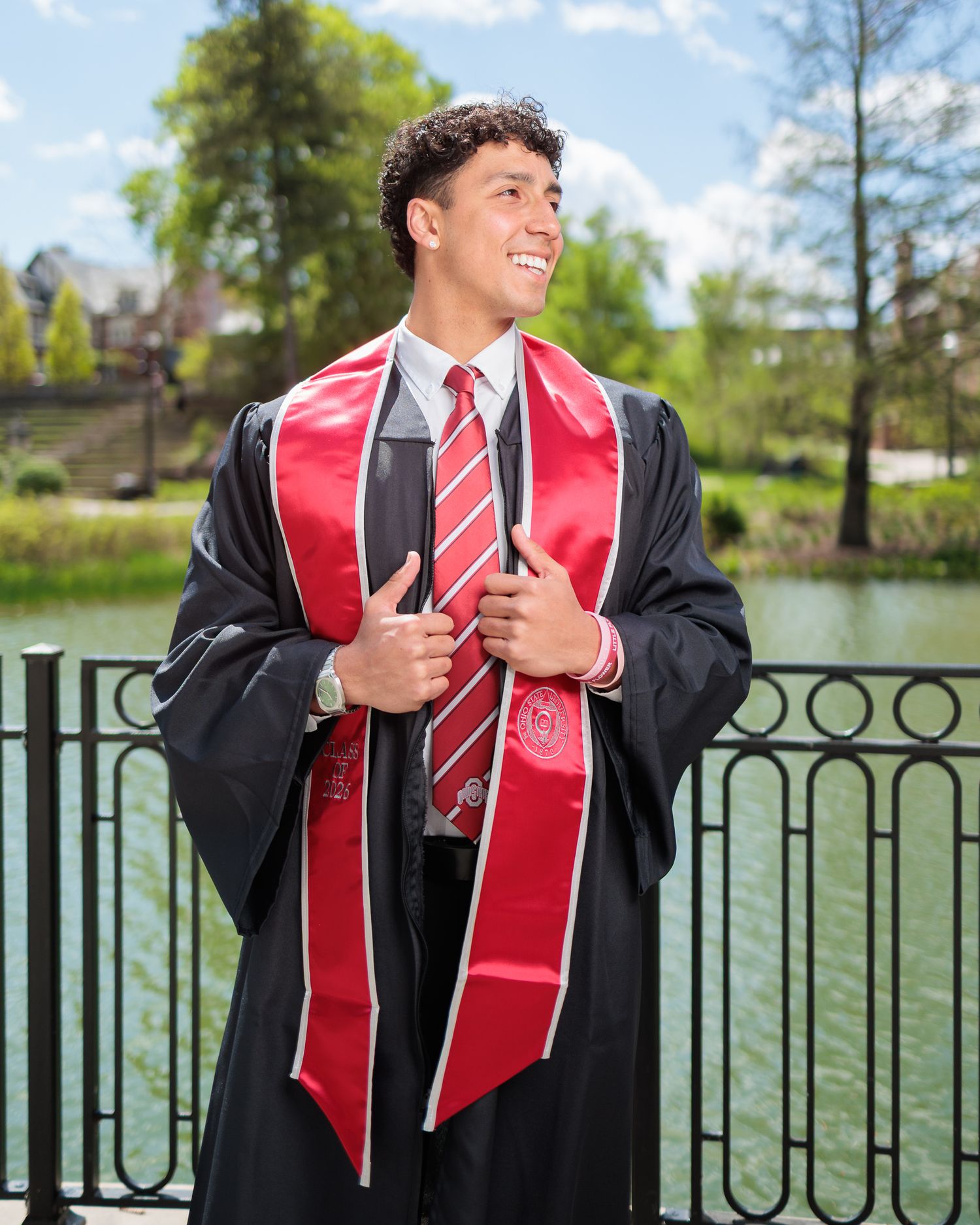 Smiling graduate in black gown and red stole poses outdoors near a pond on a sunny spring day.