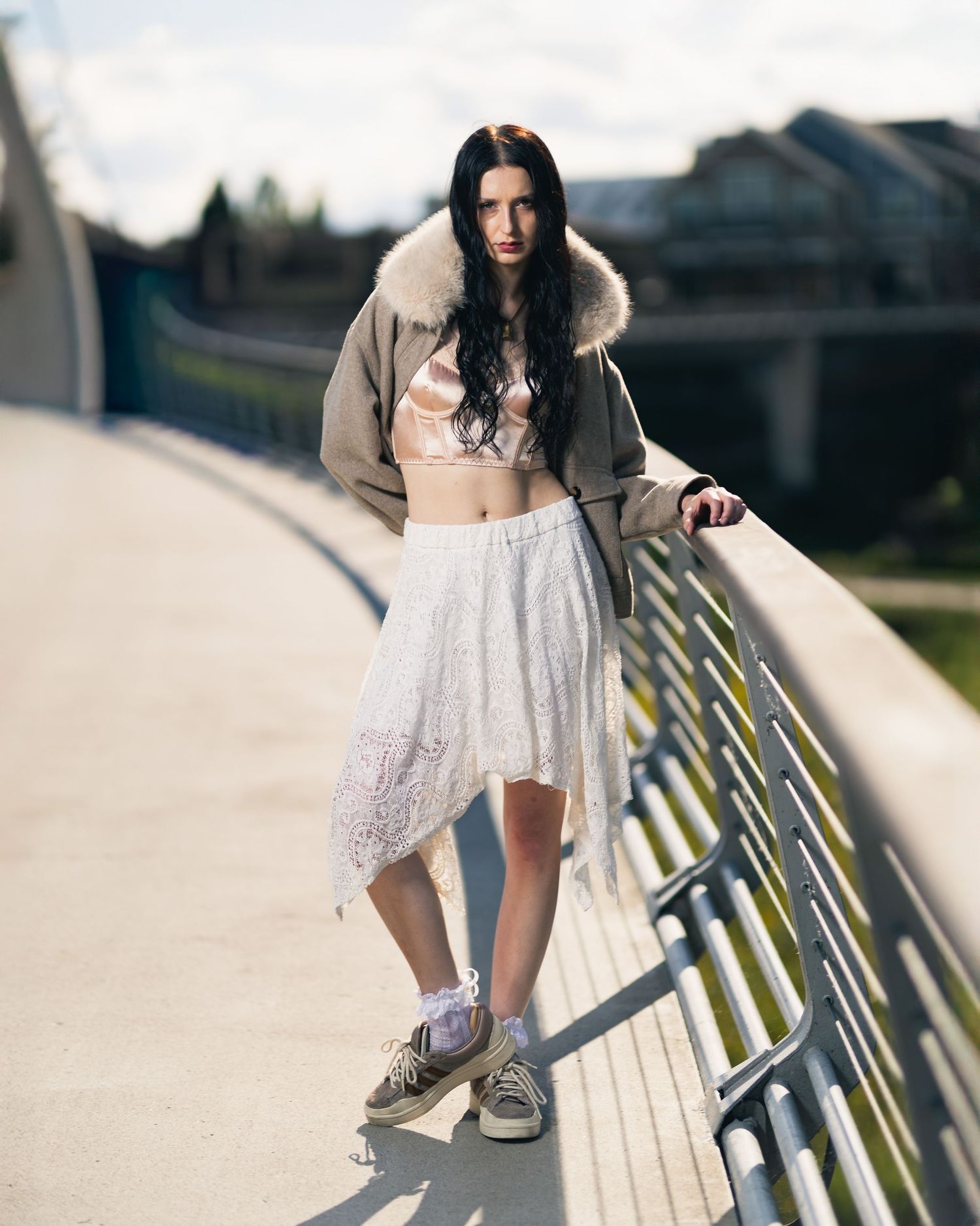 Young woman in fur-collar jacket, white lace skirt, and sneakers poses on a bridge in natural light.
