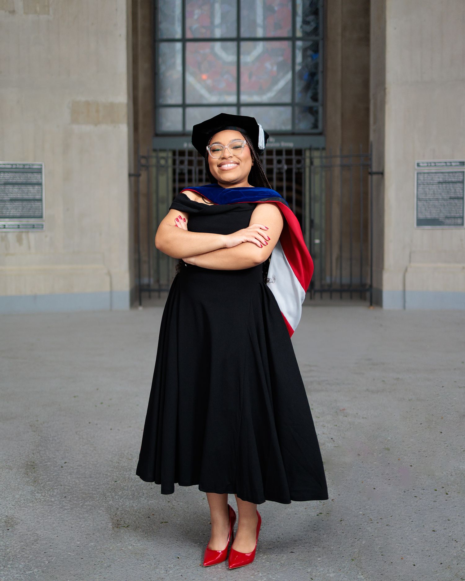 Woman in black graduation gown and cap with colorful hood, arms crossed, smiling confidently outdoors.
