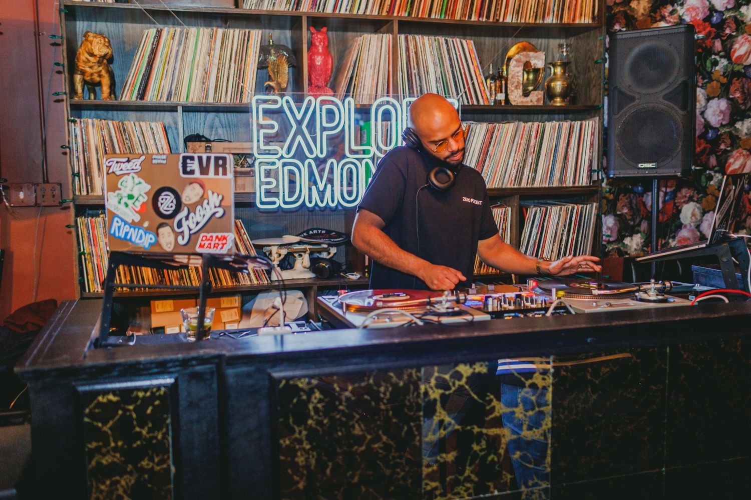 A DJ performs at a setup with vinyl records and equipment against a wall of album covers.
