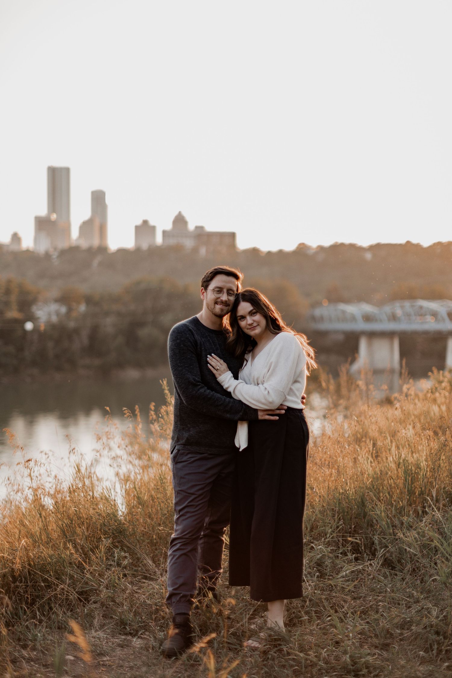 Romantic couple embraces at sunset with city skyline in background.