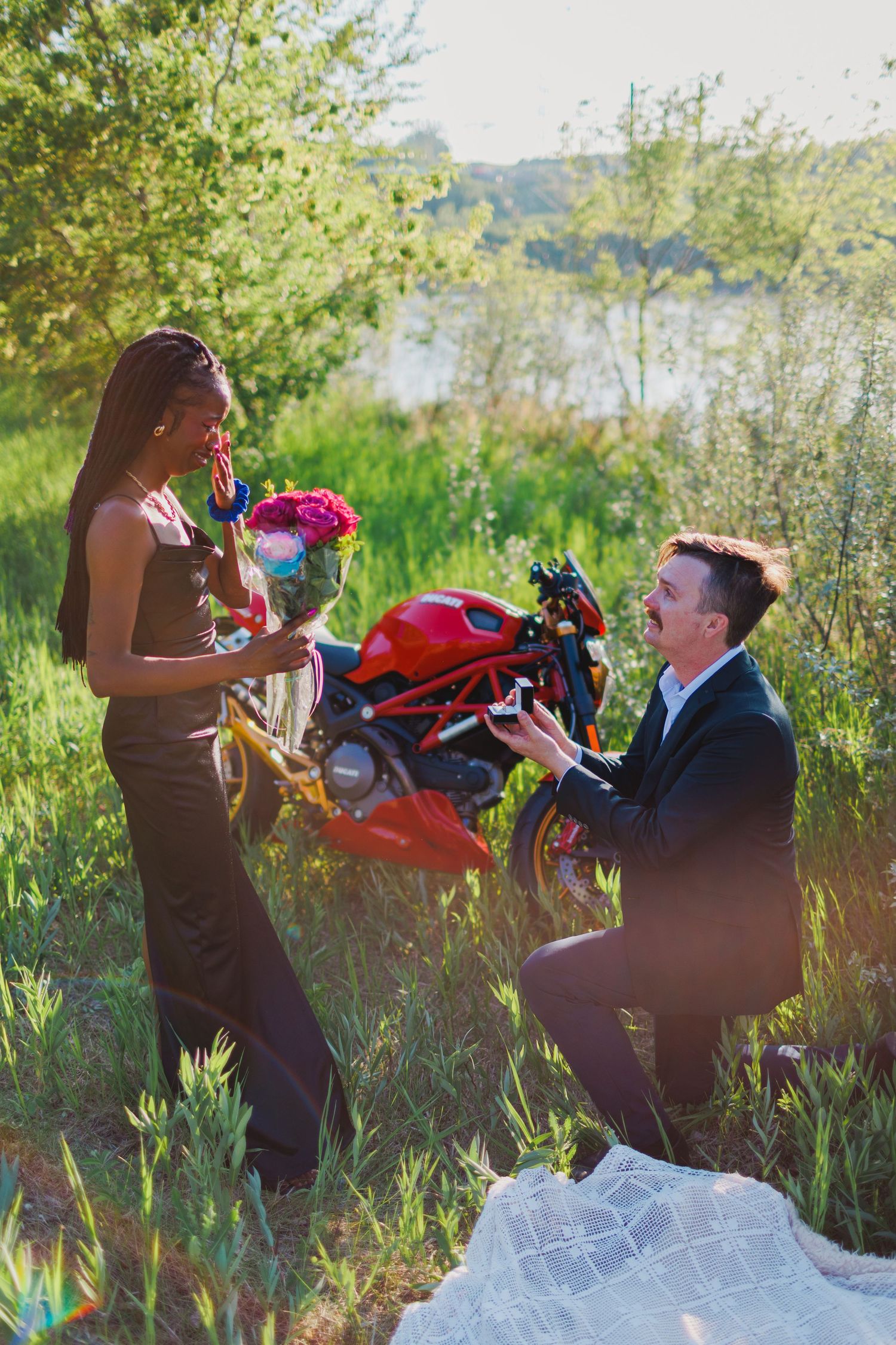 Two people in formal attire playfully tussling with red flowers in a sunlit outdoor setting with tall grass.