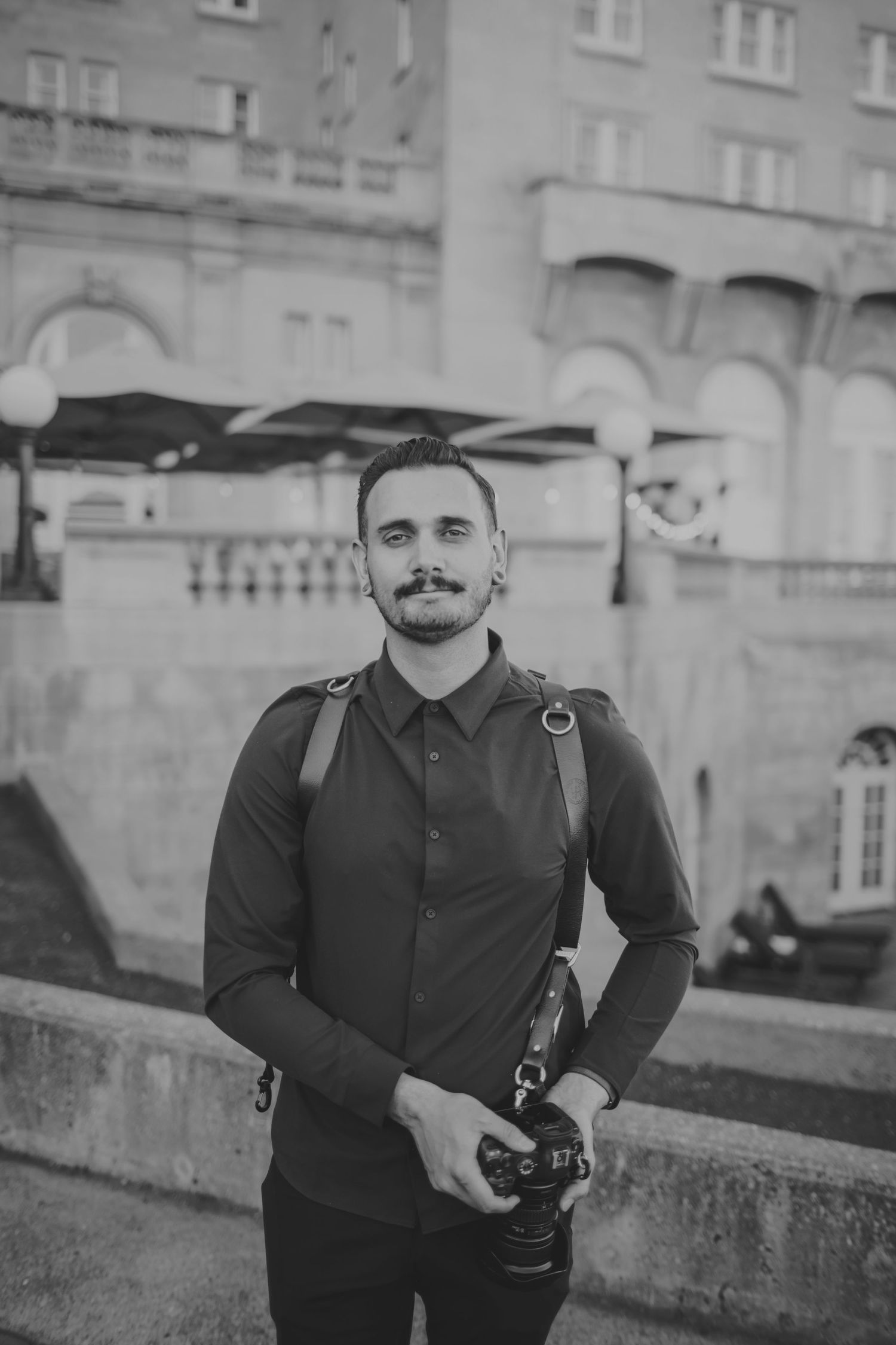 A photographer in dark clothing with a camera strap stands in front of an ornate stone building.