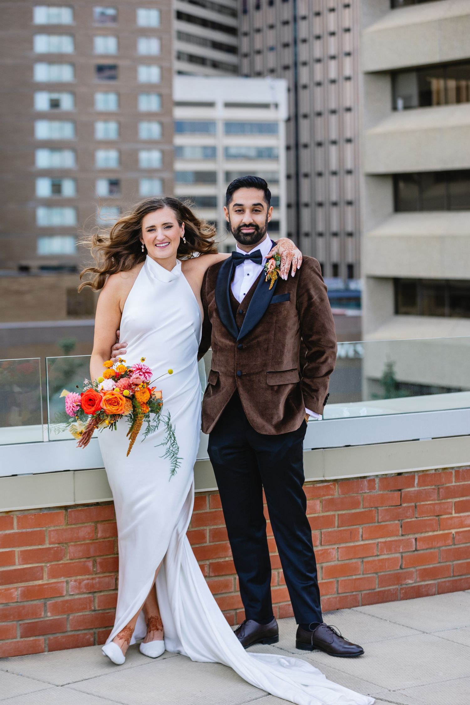 Wedding couple poses playfully on a rooftop with colorful bouquet and brick walls.