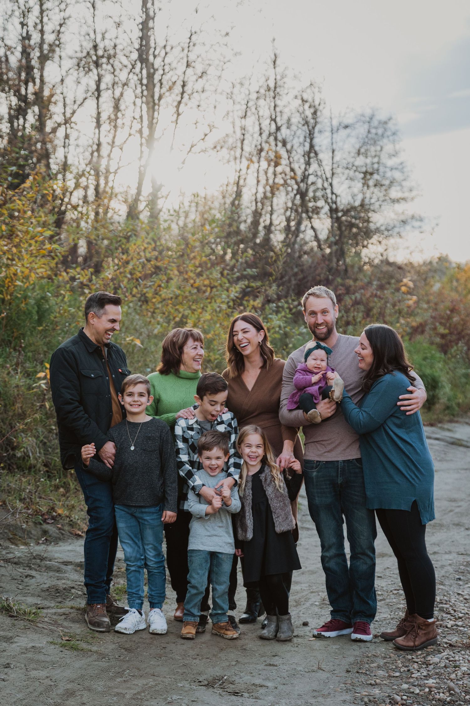 A happy family group poses together outdoors on a dirt path surrounded by autumn trees.