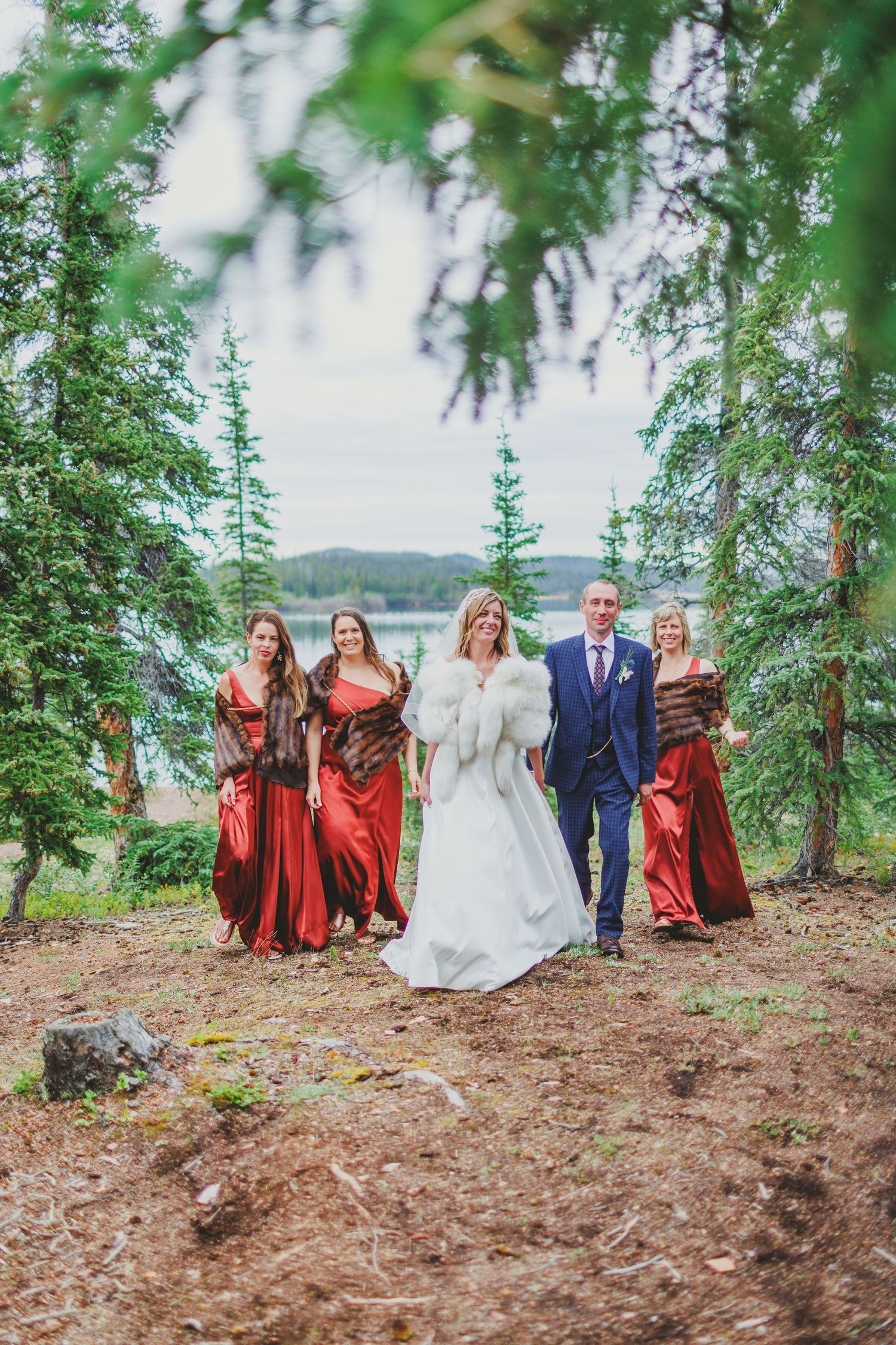 Bridal party in red dresses poses among pine trees near mountain lake.
