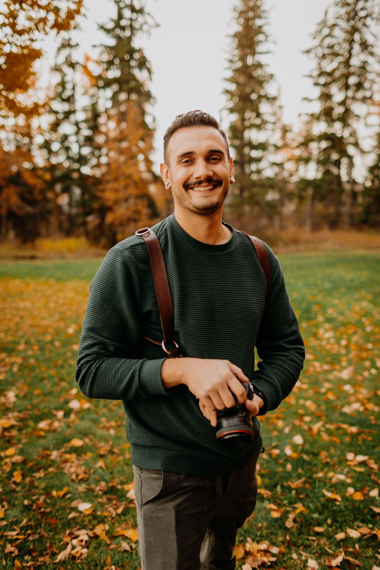 A photographer in a green sweater smiles while holding camera equipment in an autumn setting.