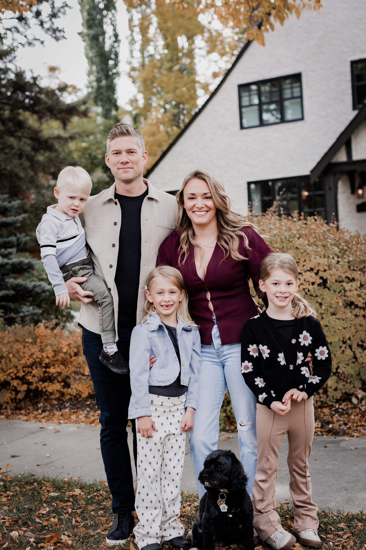 Family group poses together outdoors during fall season near white house.