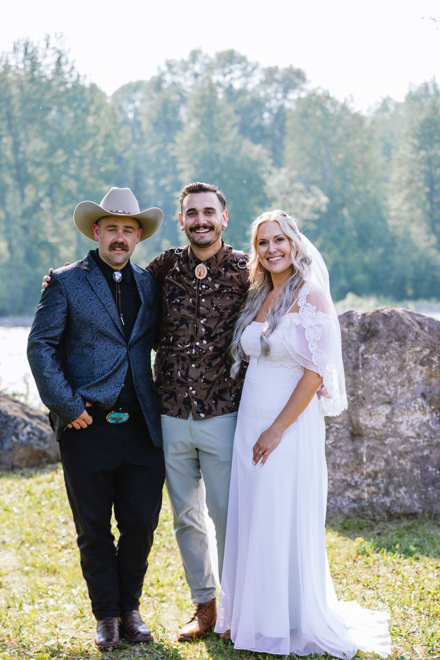 A rustic outdoor wedding scene with three people standing together near a stone wall.
