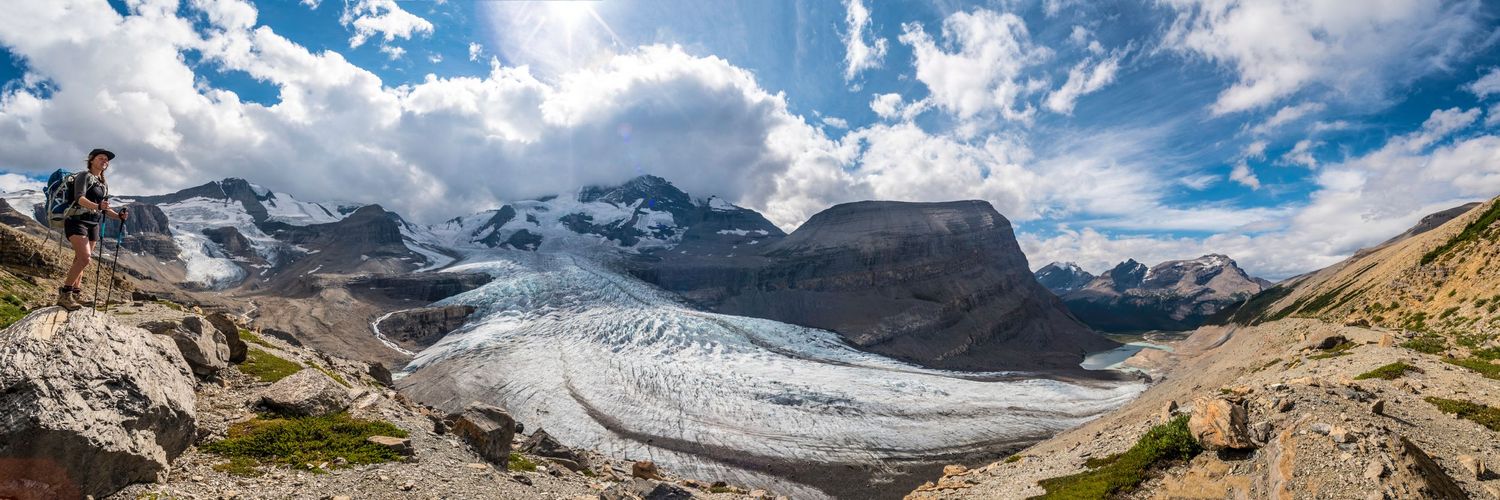 Panoramic view of a massive glacier flowing between mountain peaks under cloudy blue skies.