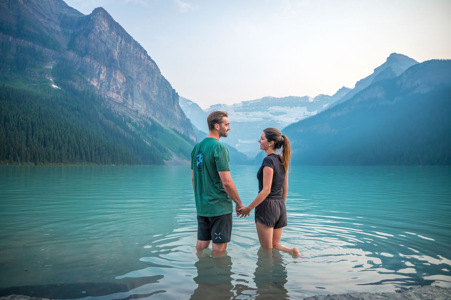 A couple standing in the tranquil turquoise waters of Lake Louise, surrounded by mountain peak,s after their engagement.