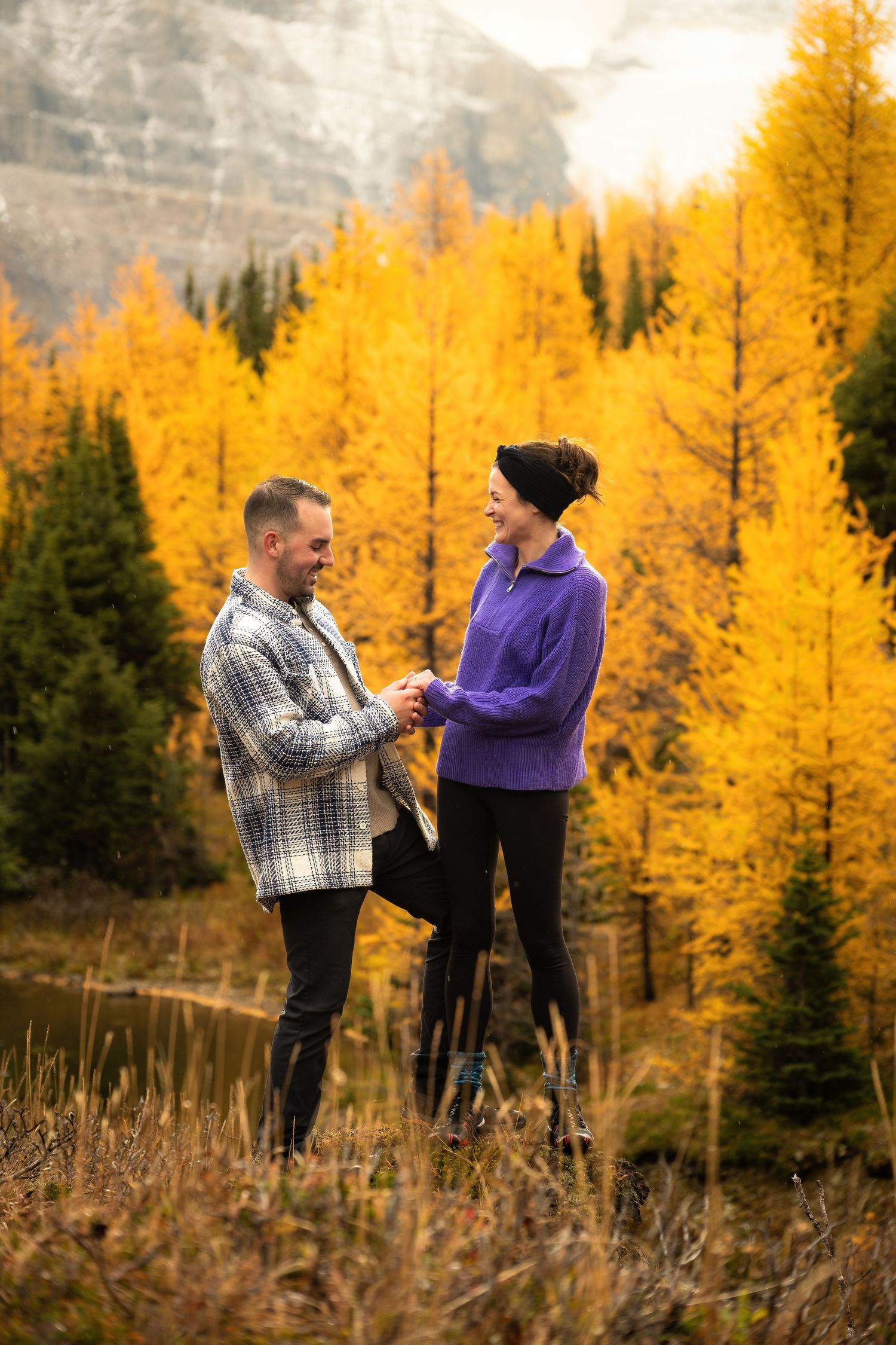 Two people stand together against a backdrop of vibrant yellow fall foliage in a mountain setting.
